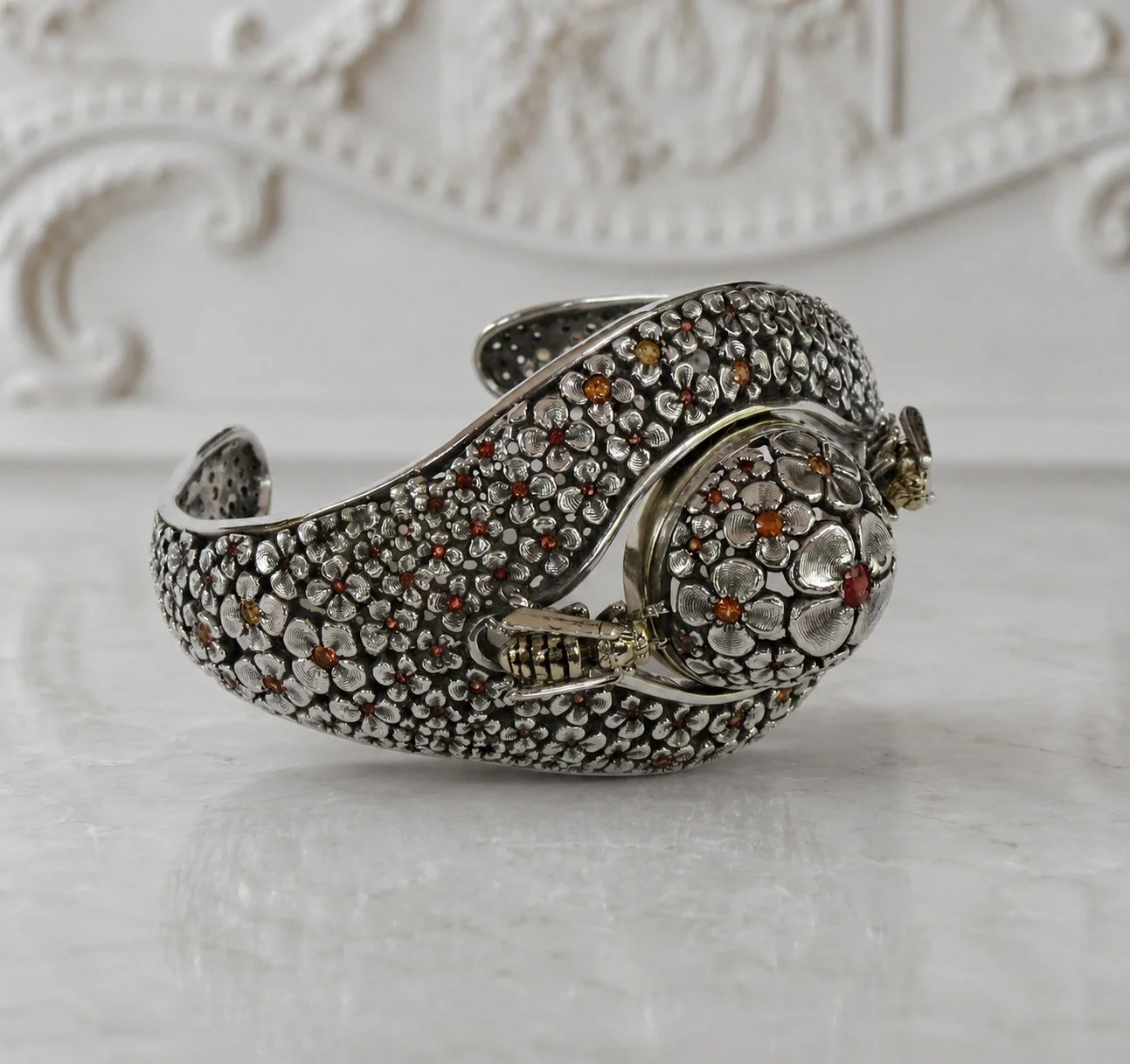A silver bracelet with intricate floral patterns and small colored gemstones, featuring a spherical clasp with matching floral design and gemstones, resting on a white surface with a decorative white background.