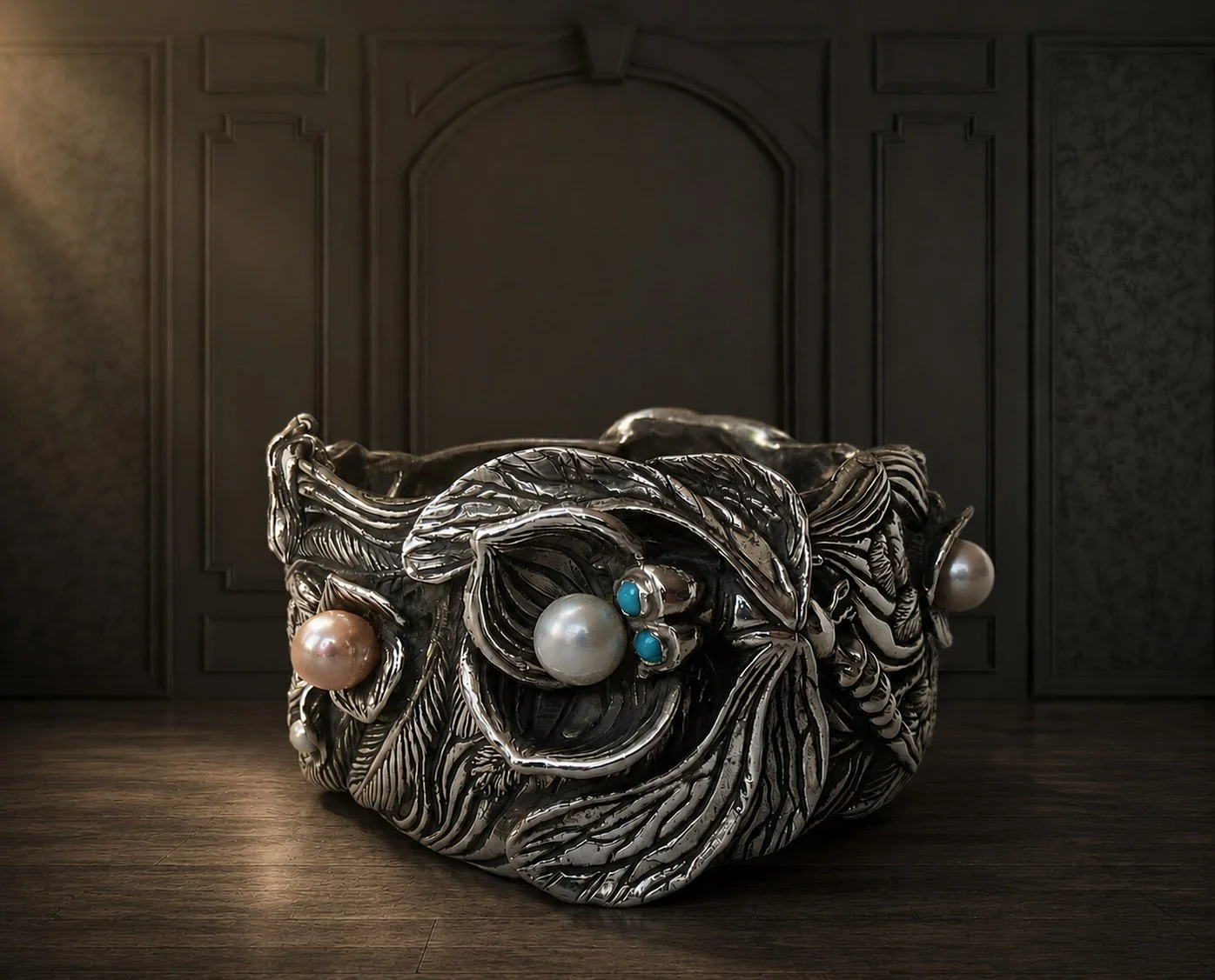 A decorative, ornate silver bowl with intricate leaf and floral patterns, adorned with large pearls and turquoise embellishments, set on a wooden floor against a dark, paneled wall.