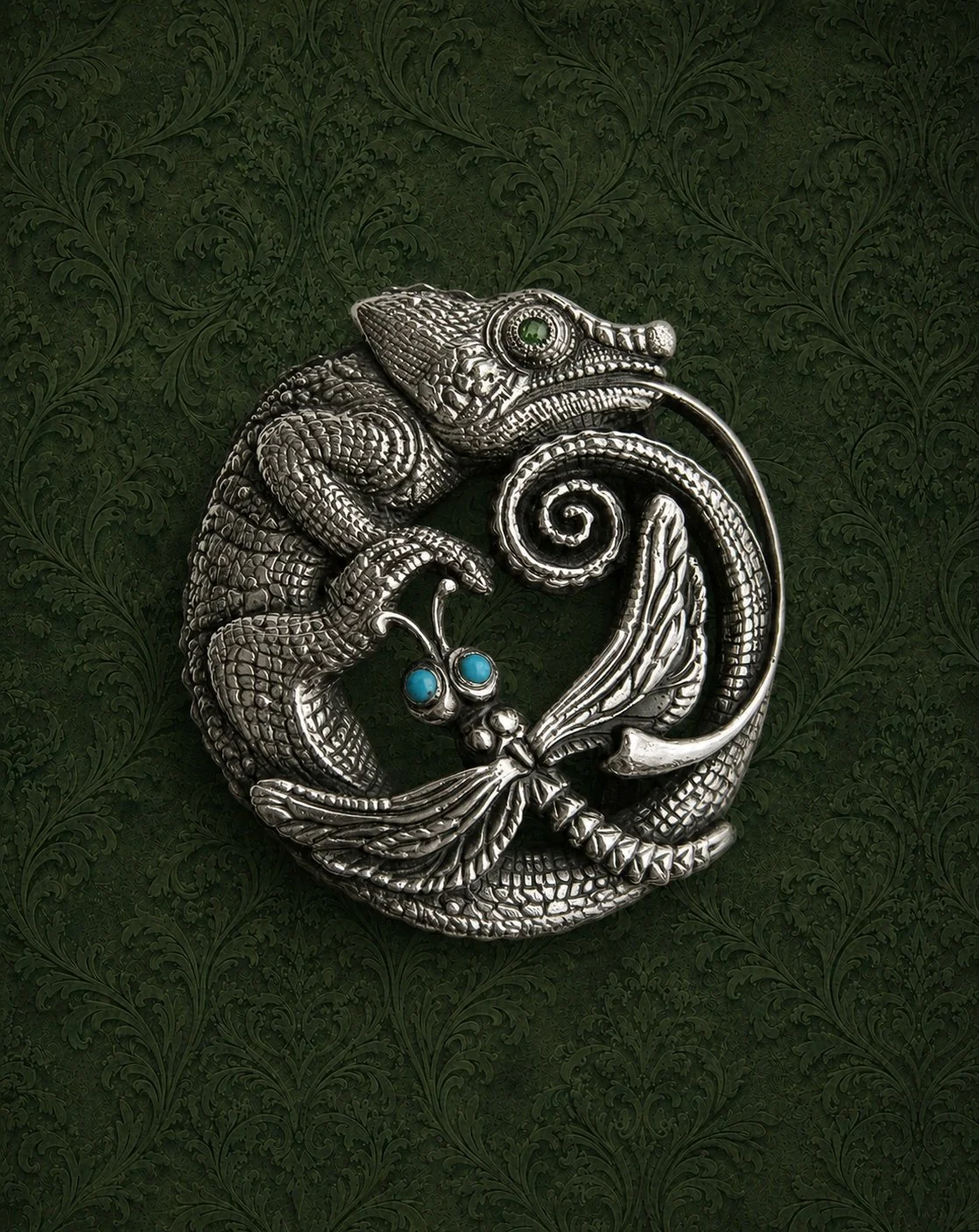 A silver chameleon-shaped pin with green and blue gemstone eyes, resting on a dark green textured background.