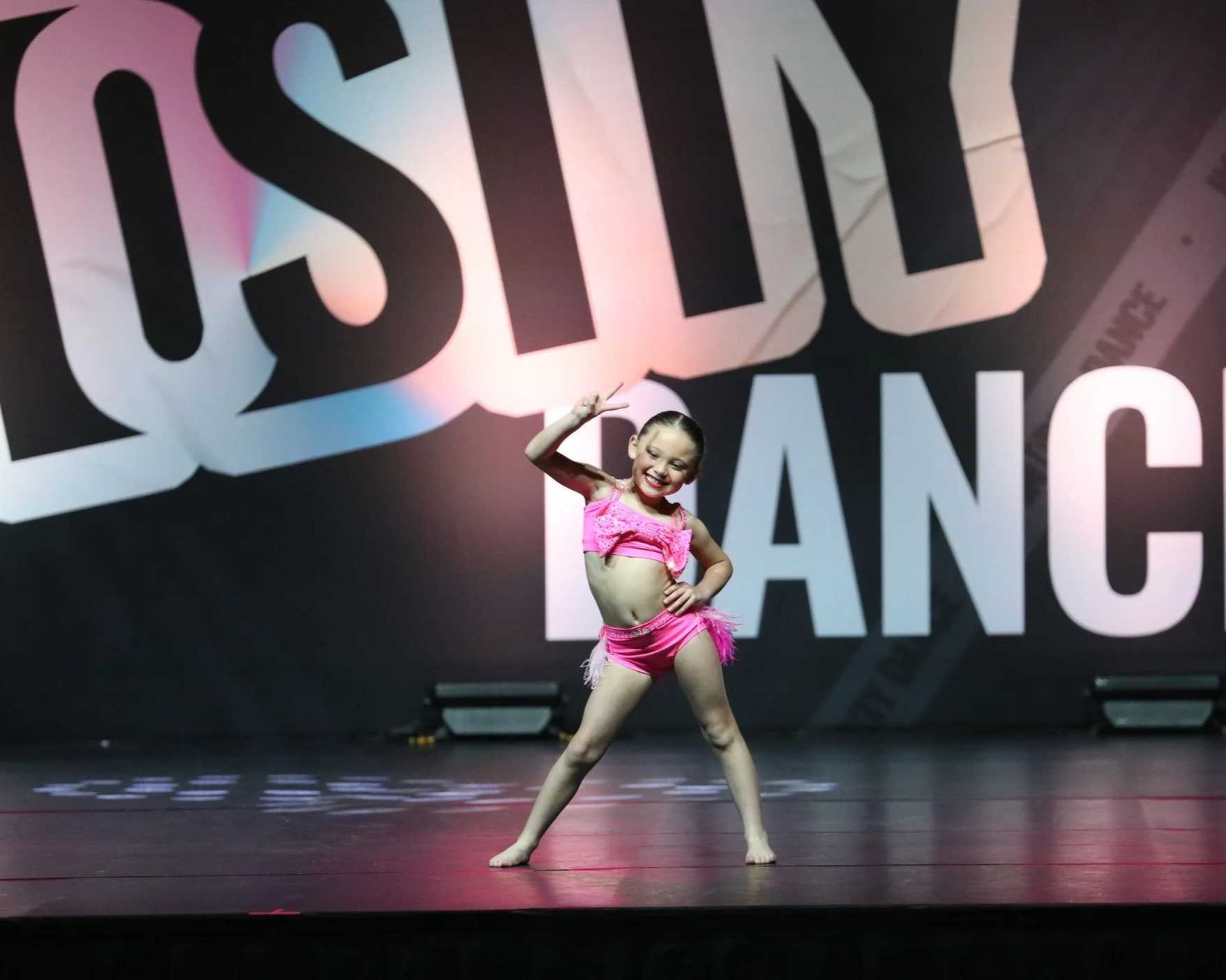 Young girl in a pink dance costume striking a pose on stage with a large sign in the background.