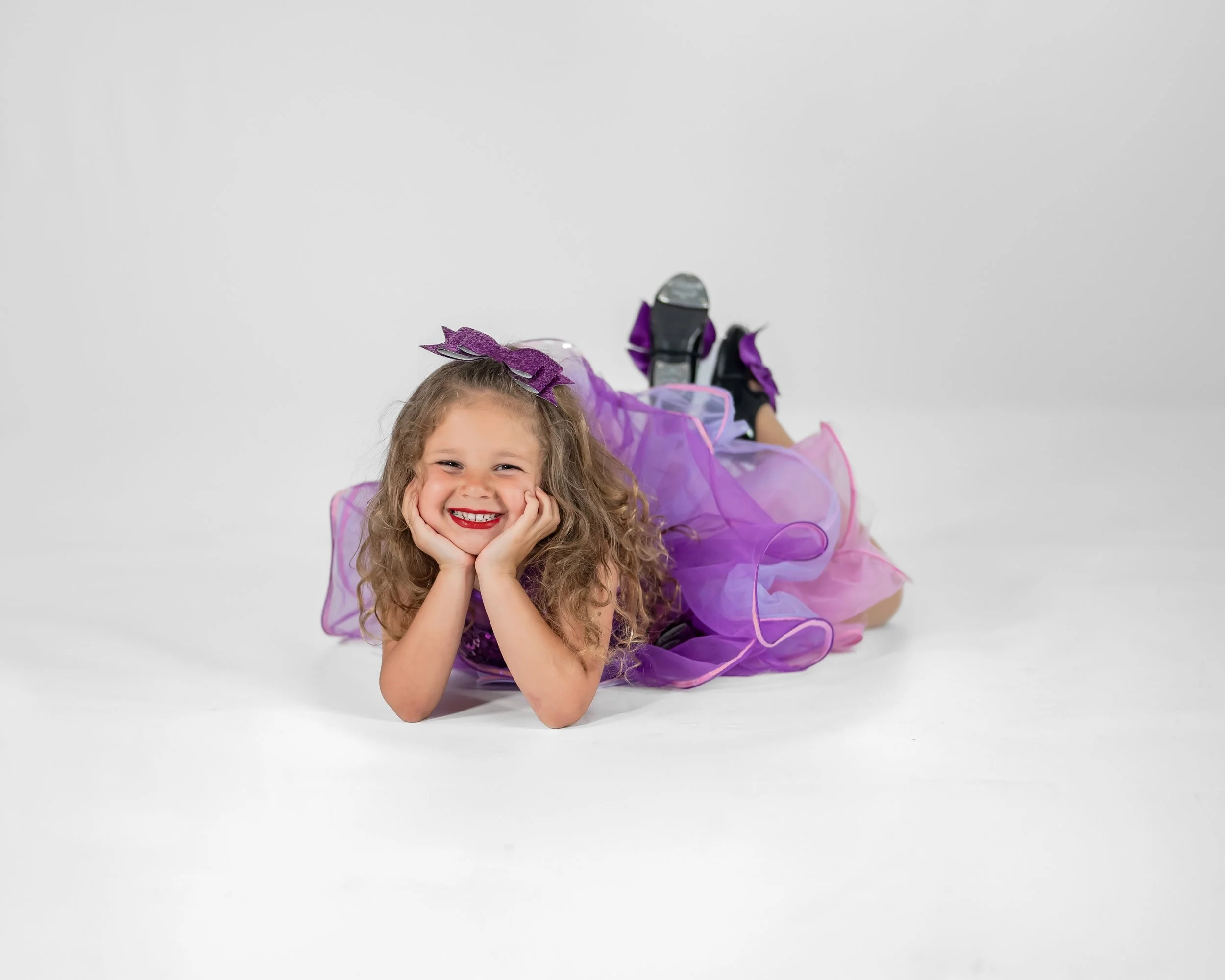 A young girl lying on her stomach, smiling with her chin resting on her hands, wearing a purple fairy costume with a tutu and sparkly headband, against a plain white background.