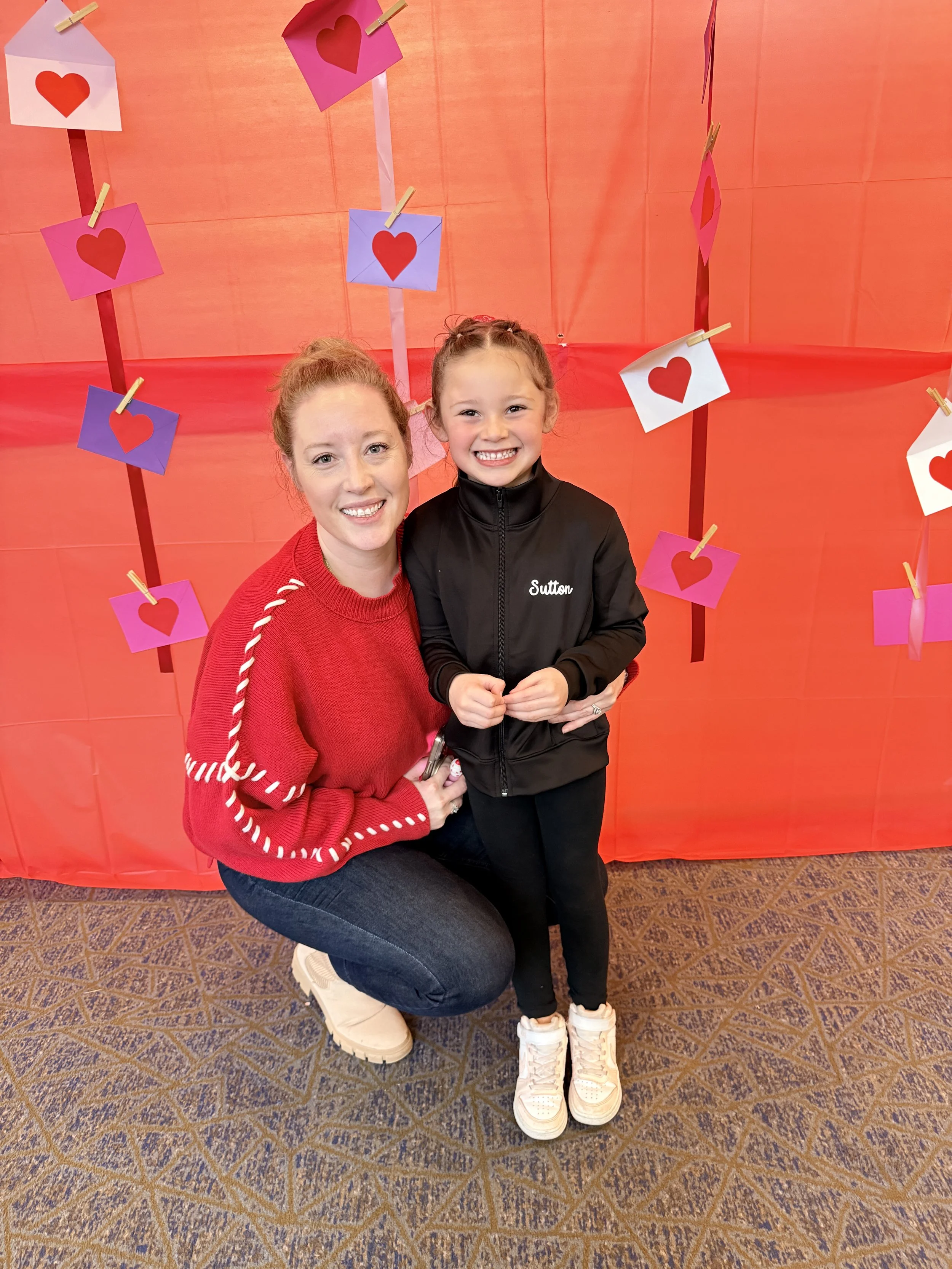 A woman and a young girl smiling at the camera, standing in front of a red backdrop decorated with paper hearts and colorful cards attached with clothespins.