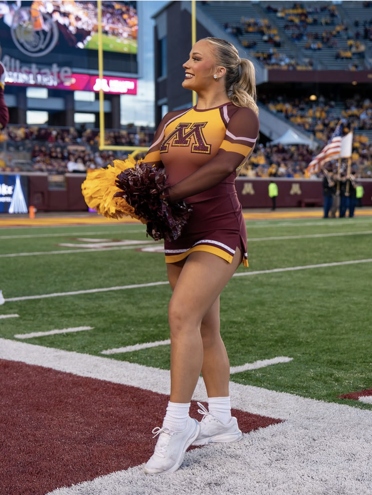 A smiling cheerleader in maroon and gold uniform holding pom-poms on a football field before a game, with stadium and crowd in the background.