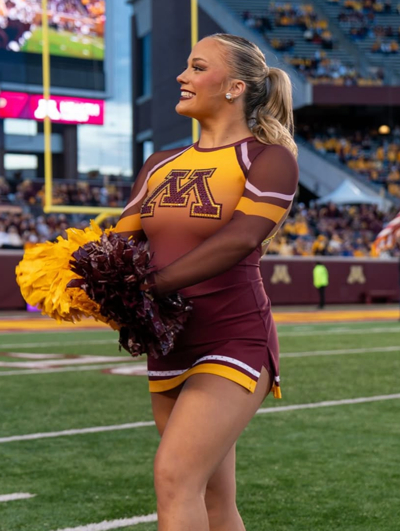 A smiling cheerleader in a maroon and gold uniform with a large letter 'M' on her top, holding yellow and maroon pom-poms on a football field during a game.