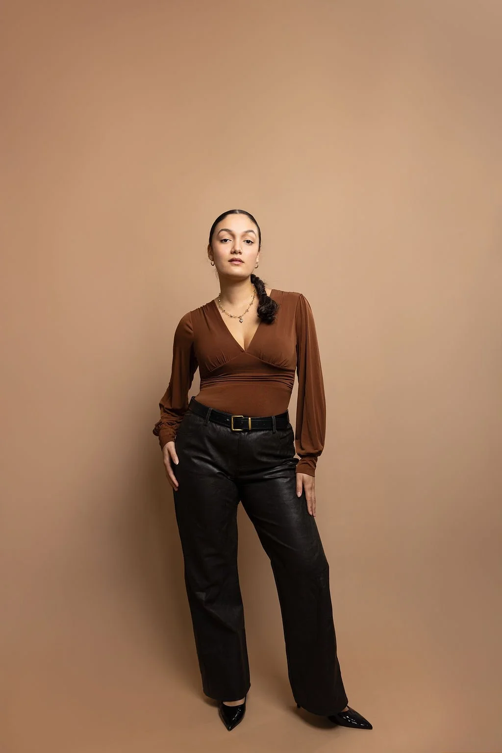 A woman standing against a beige background, wearing a brown long-sleeve top, black pants, and black high heels, with her right hand in her pocket and a confident expression.