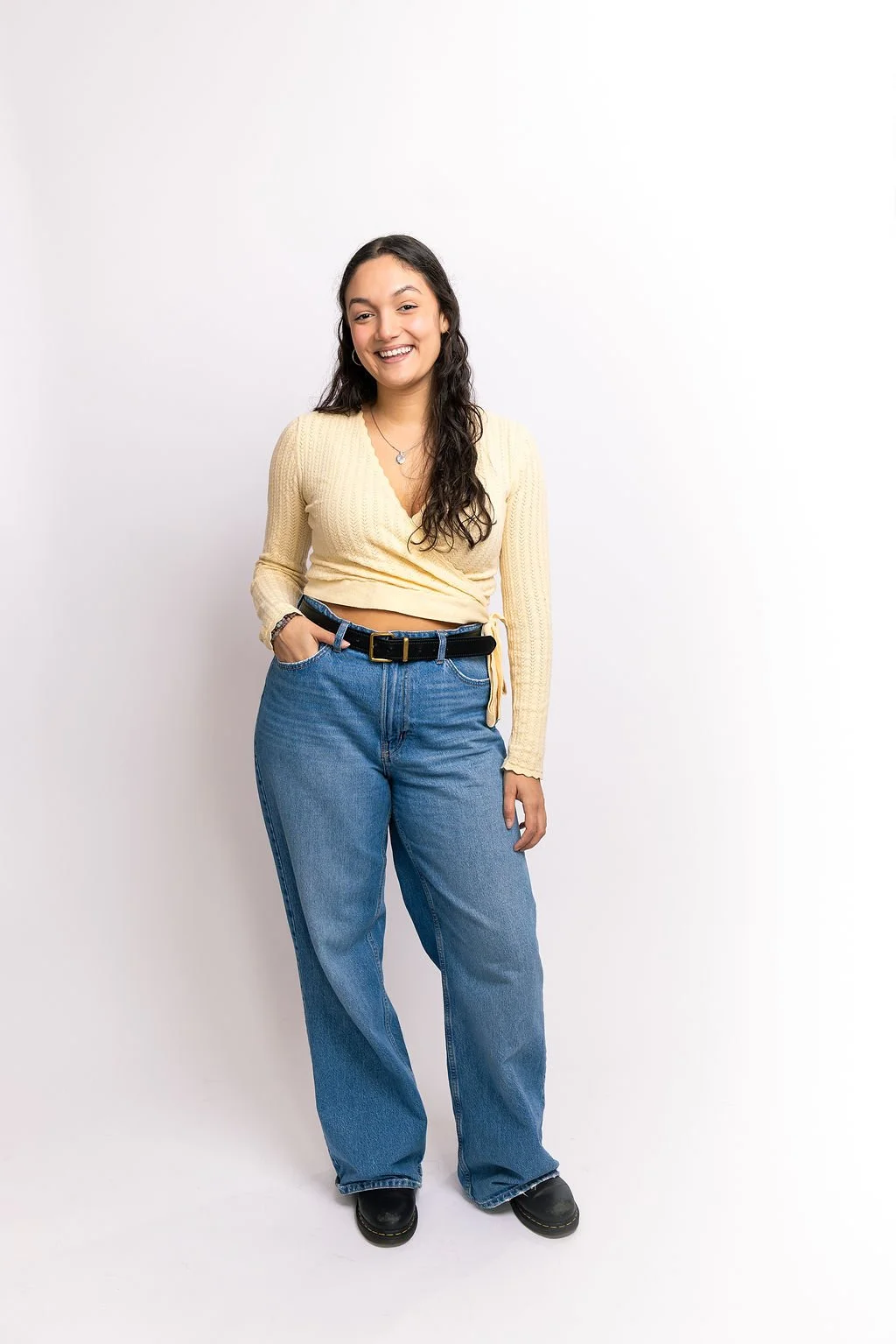 Young woman with long, dark wavy hair, smiling, wearing a yellow cropped sweater, blue jeans, and black boots, standing against a plain white background.