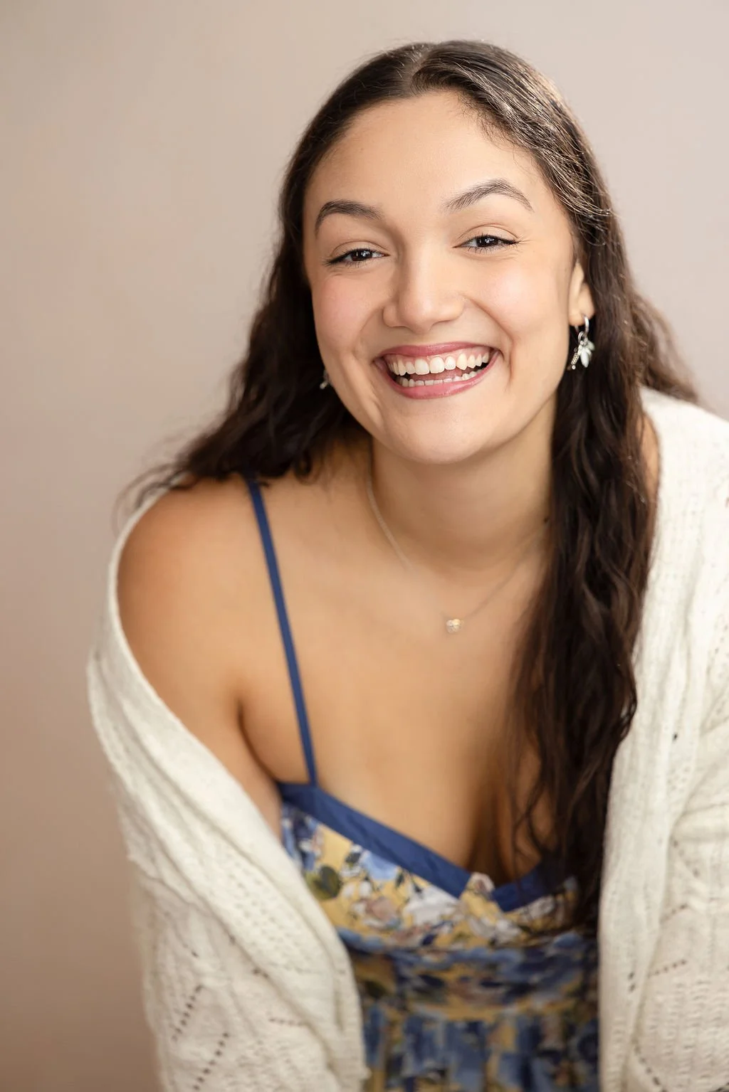 A young woman with long dark hair smiling and looking at the camera. She is wearing earrings, a necklace, a blue floral top, and a cream-colored cardigan.
