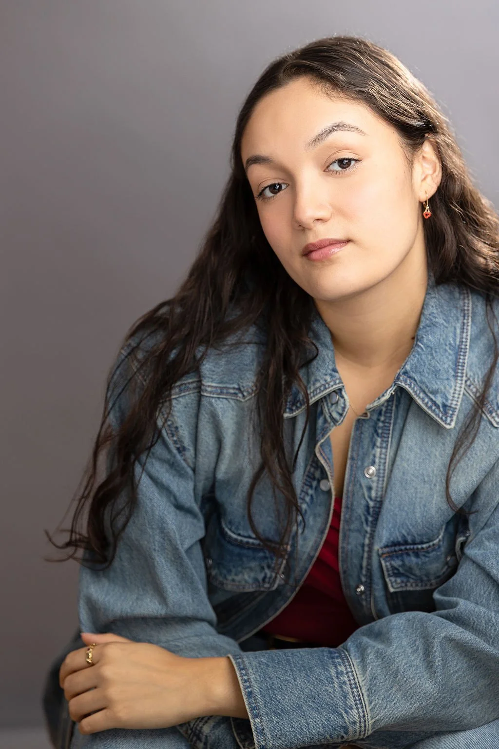 A young woman with long curly dark hair wearing a denim jacket and a red top, looking at the camera with a neutral expression, posing against a plain gray background.