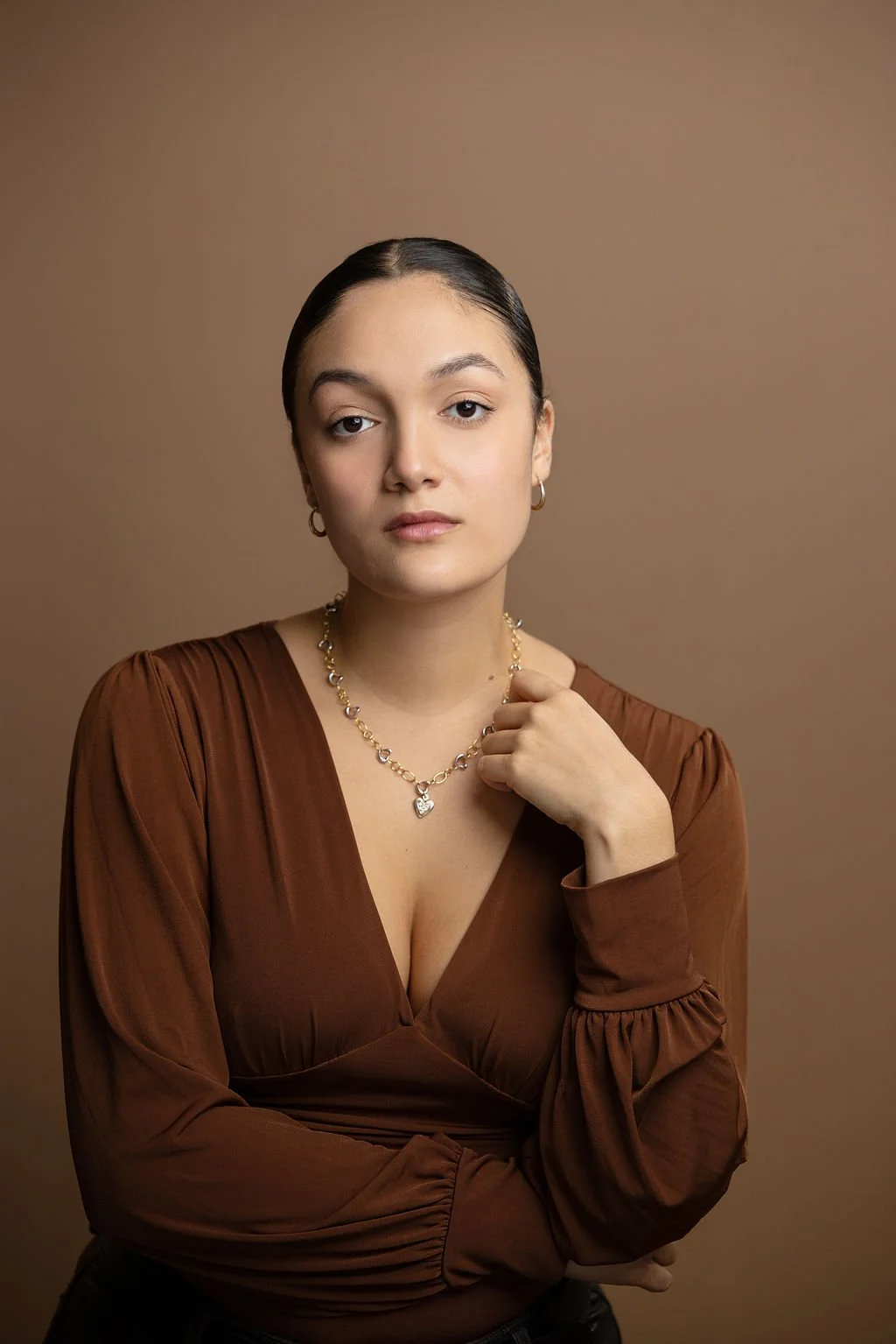 Portrait of a woman with dark hair pulled back, wearing gold jewelry and a brown top with a deep neckline, against a brown background.