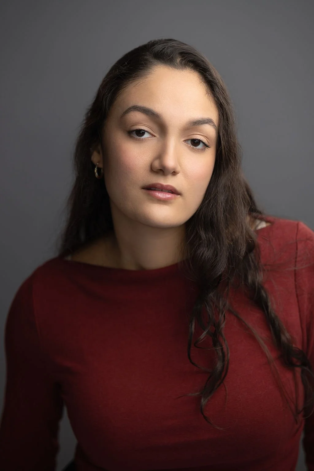A young woman with long, wavy brown hair and gold hoop earrings, wearing a maroon top, looking at the camera with a neutral expression against a gray background.