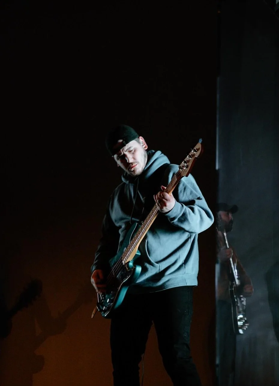 A man wearing a gray hoodie, black cap, and black pants is playing an electric bass guitar on stage, with dark lighting and smoke around him.