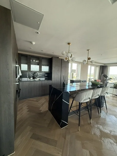 Modern open-concept kitchen with black marble island, gray cabinets, and pendant lighting, adjacent to a living area with large windows.