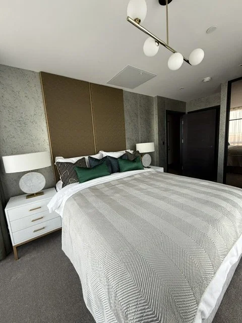 A modern bedroom with a large bed, white nightstands with lamps, a gold and textured accent wall behind the bed, and a contemporary hanging light fixture with spherical bulbs.