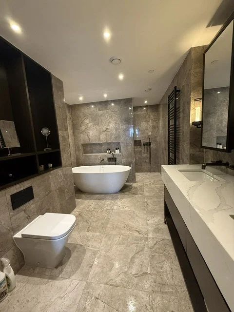 Modern bathroom with a white freestanding bathtub, a wall-mounted toilet, a large marble sink with a mirror, and gray tiled walls and floors.