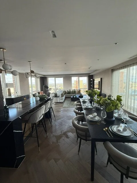 Modern open-concept living and dining room with a long black dining table set with plates and glassware, surrounded by beige chairs, with a spacious white sofa area and large windows allowing natural light.