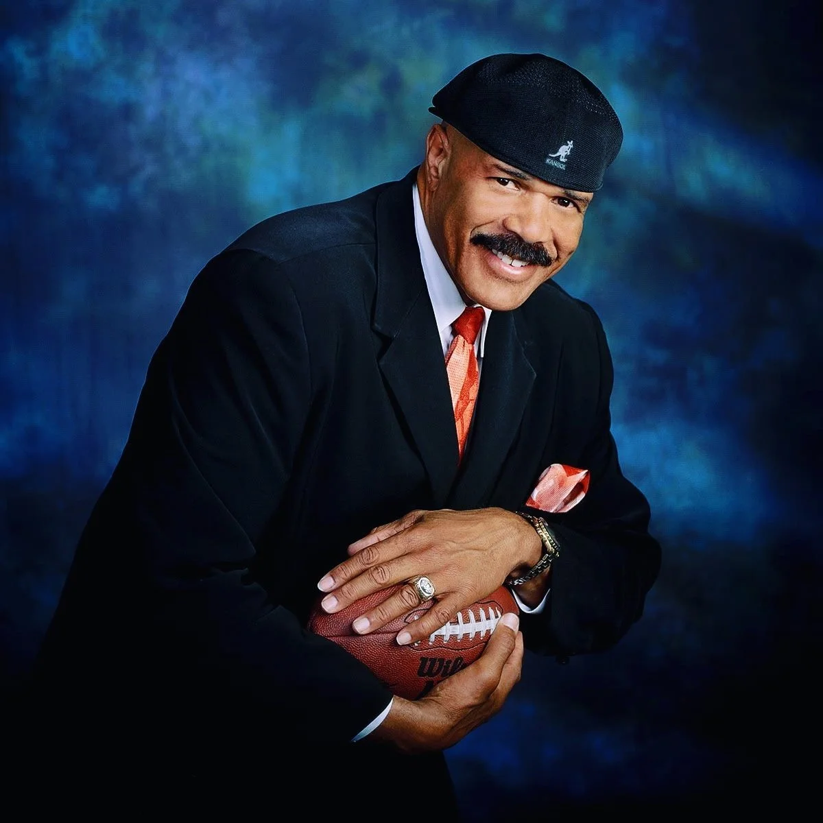 A man in a black suit, white shirt, and orange tie holding an American football, wearing a black cap with a logo, smiling at the camera against a blue textured background.