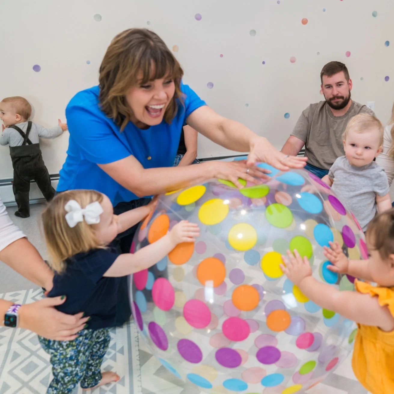 Children and adults playing with a large, colorful, polka-dotted beach ball in a room with a white wall and pastel polka-dot decorations.