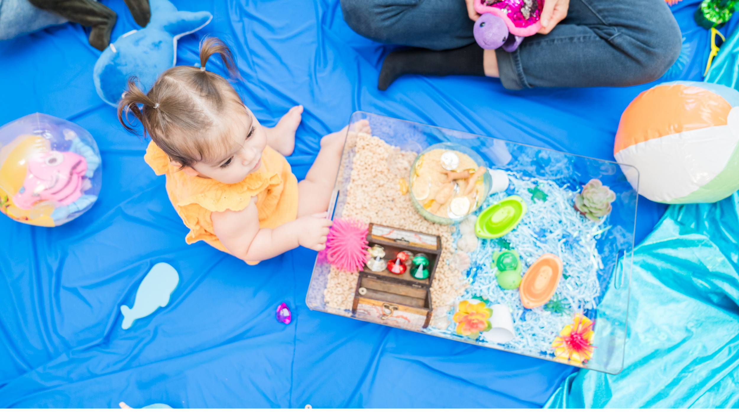 A young girl in an orange dress sitting on a blue beach mat surrounded by colorful toys and decorations, including a box of small toys, balloons, and plush sea creatures, during a playful outdoor gathering.