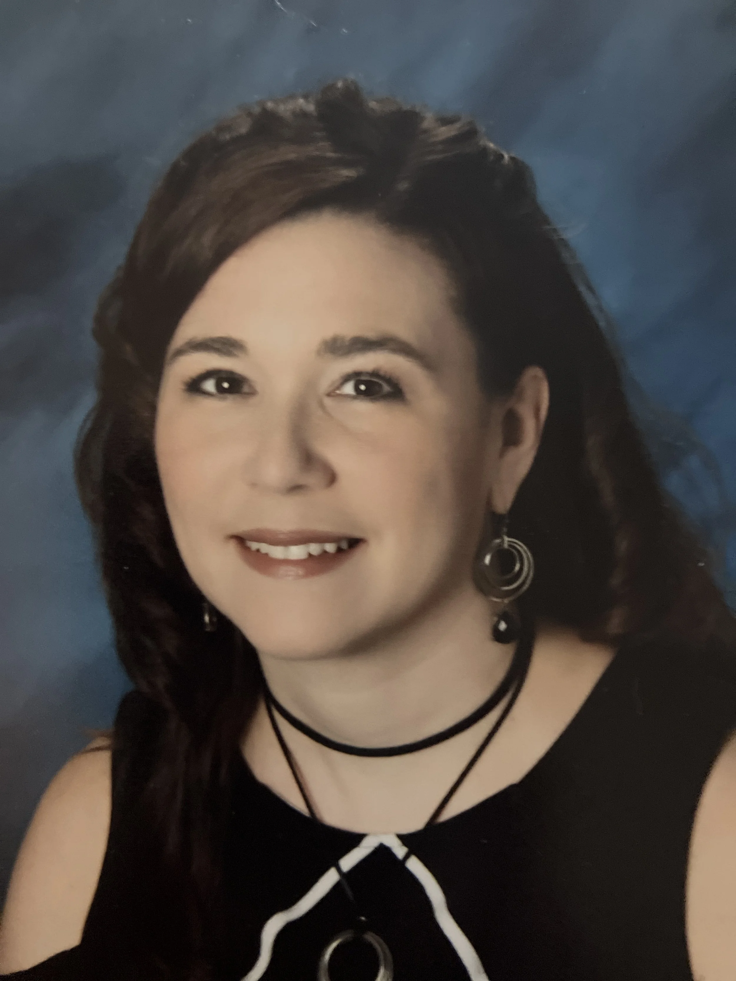 Close-up portrait of a woman with brown hair, smiling, wearing black earrings, necklaces, and a black top with a white outline design.