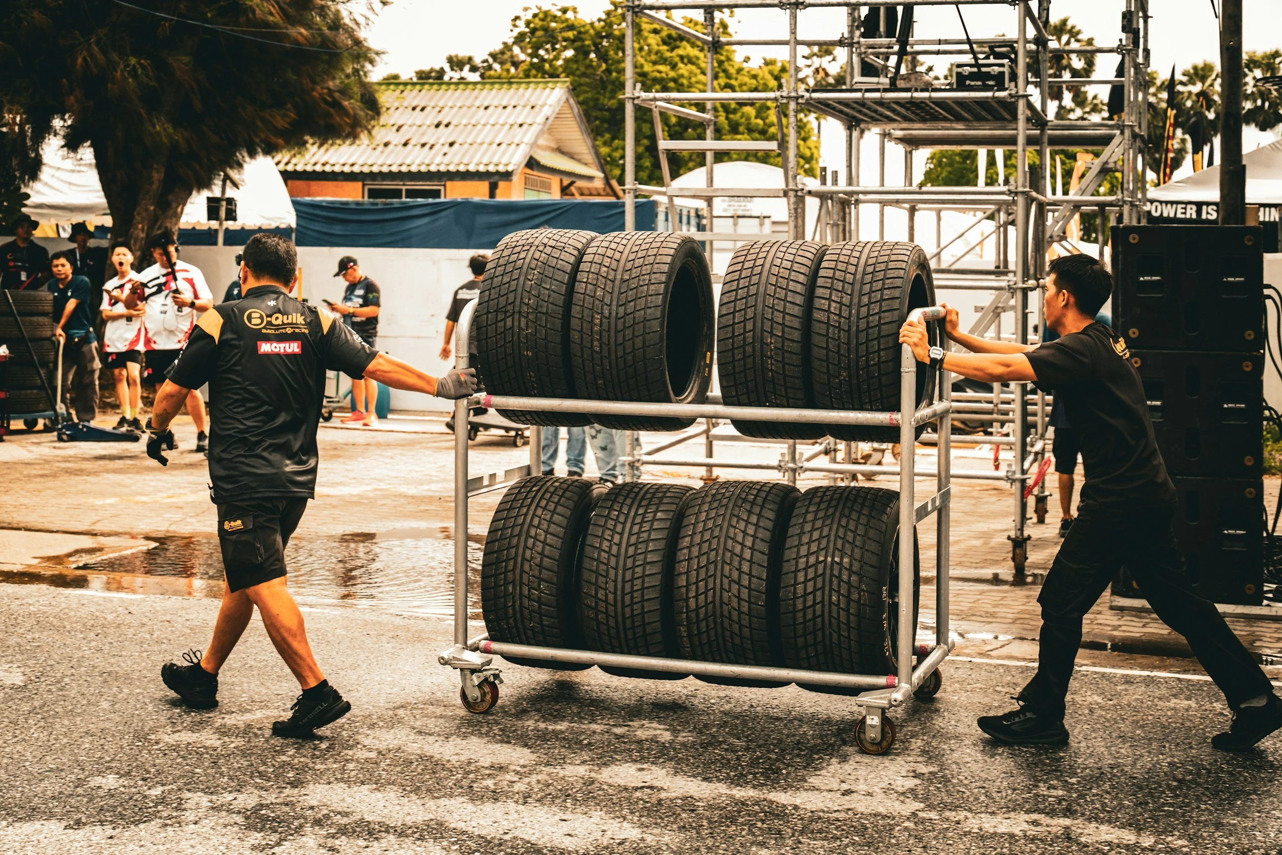 Two people are moving a large metal cart with stacked tires at an outdoor event, possibly a tire change or race preparation. There are several onlookers and a scaffolding structure in the background.