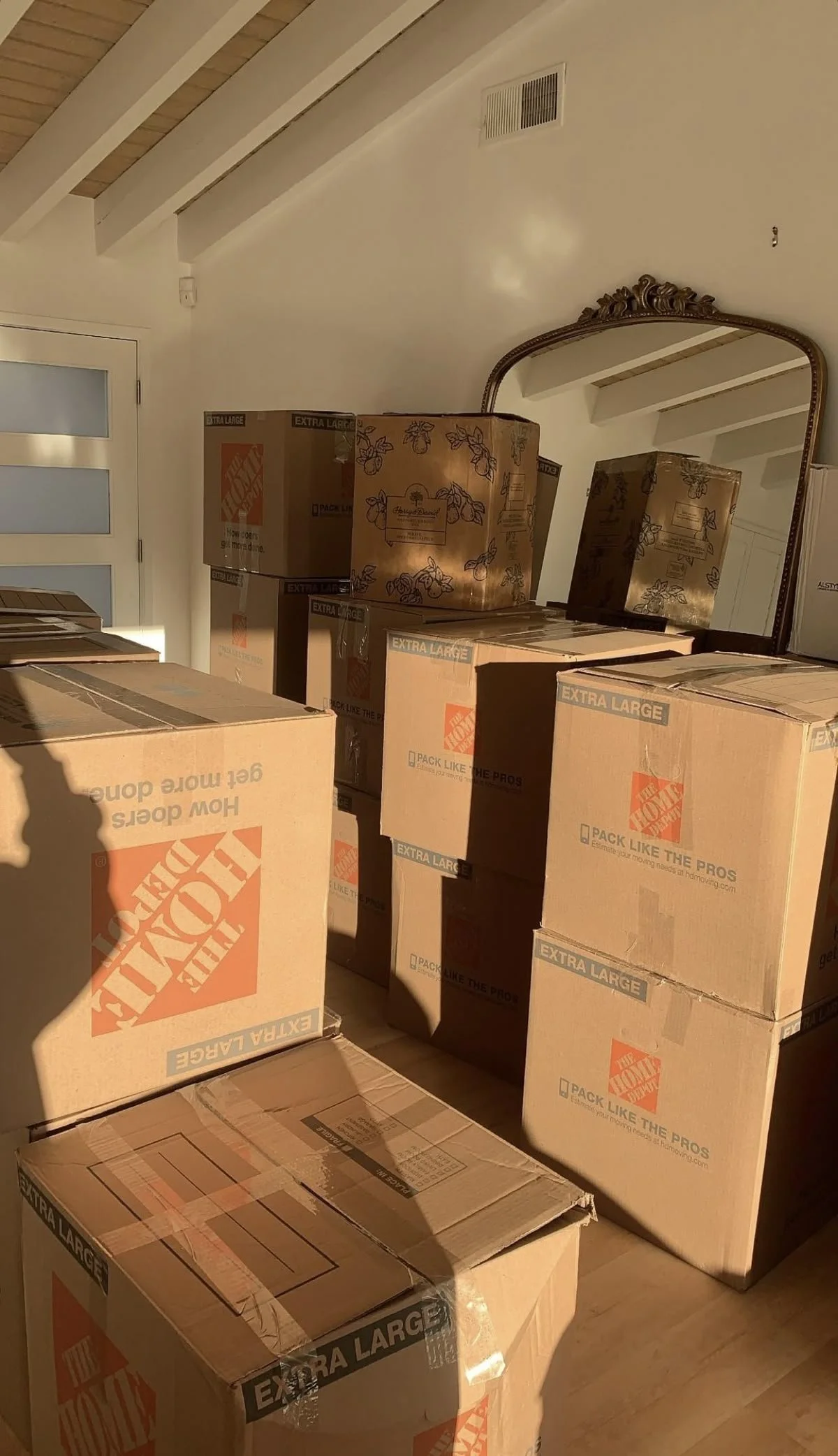 Moving boxes stacked on a wooden floor in a room with a ceiling vent and a large ornate mirror reflecting part of the room and boxes.