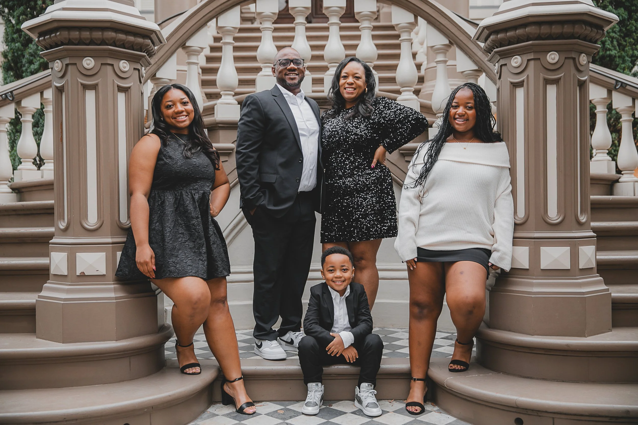 Malonn Barnes  family of five celebrating on the stairs of a historic building. Four women, one man, and a young boy are dressed in formal and semi-formal attire, smiling for the photo. The women wear dresses, and the man and boy wear suits.