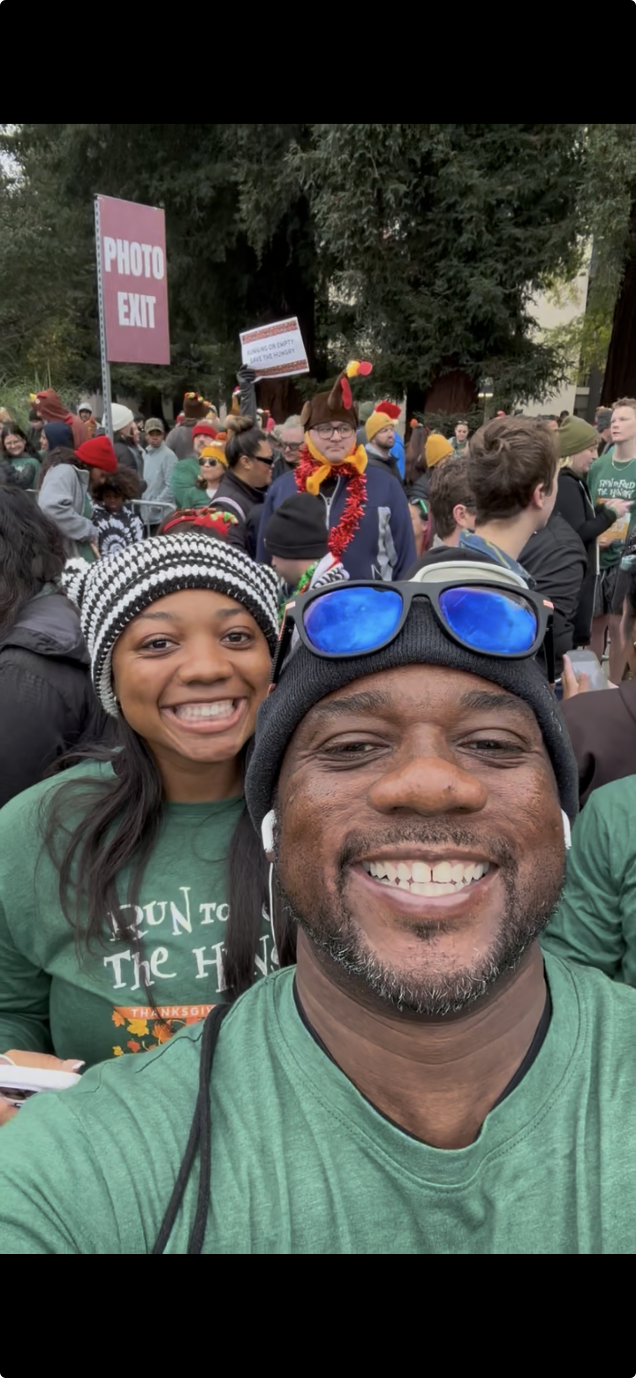 Malonn And daughter Two staking a selfie at a large outdoor gathering, with many people in Thanksgiving-themed clothing and turkey hats in the background. For Feed the HUNGRY RUN