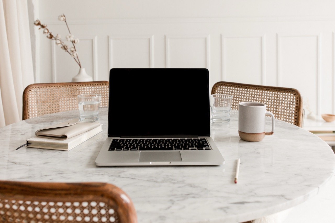 A laptop, a notebook, a pen, a mug, two glasses of water, and a vase with branches on a white marble table in a bright room.