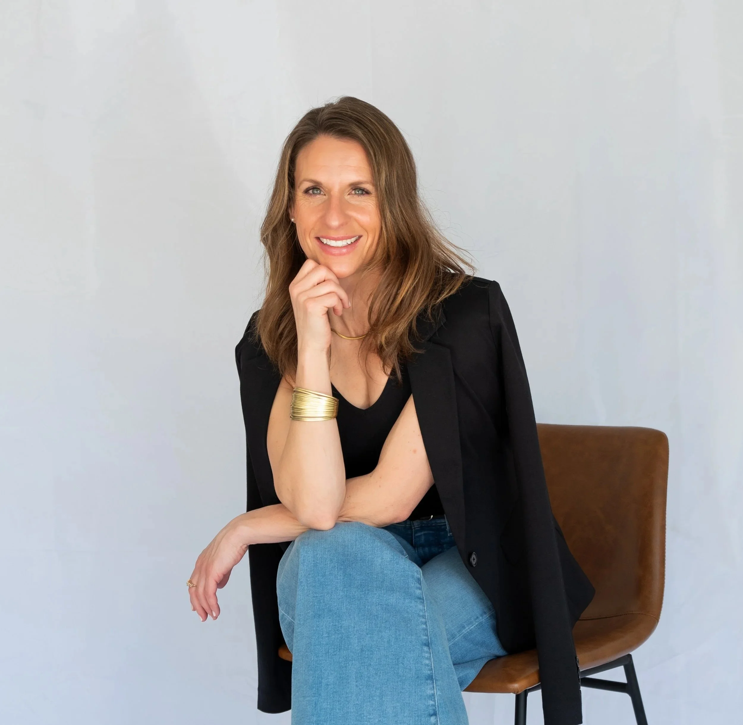 A woman with long brown hair, wearing a black top, gold jewelry, and blue jeans, sitting on a chair with her chin resting on her hand in front of a plain light wall.