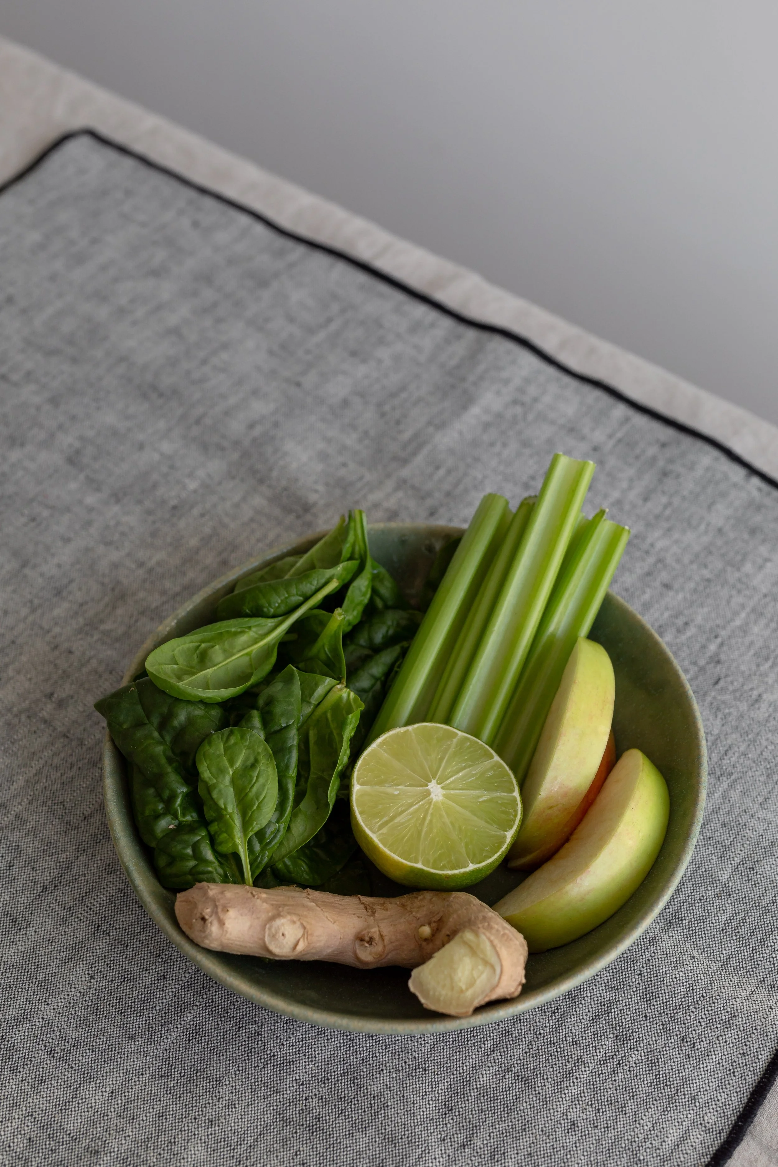 A bowl of fresh vegetables including spinach, celery, a lime half, apples, ginger, and a slice of lemon, placed on a gray fabric surface.