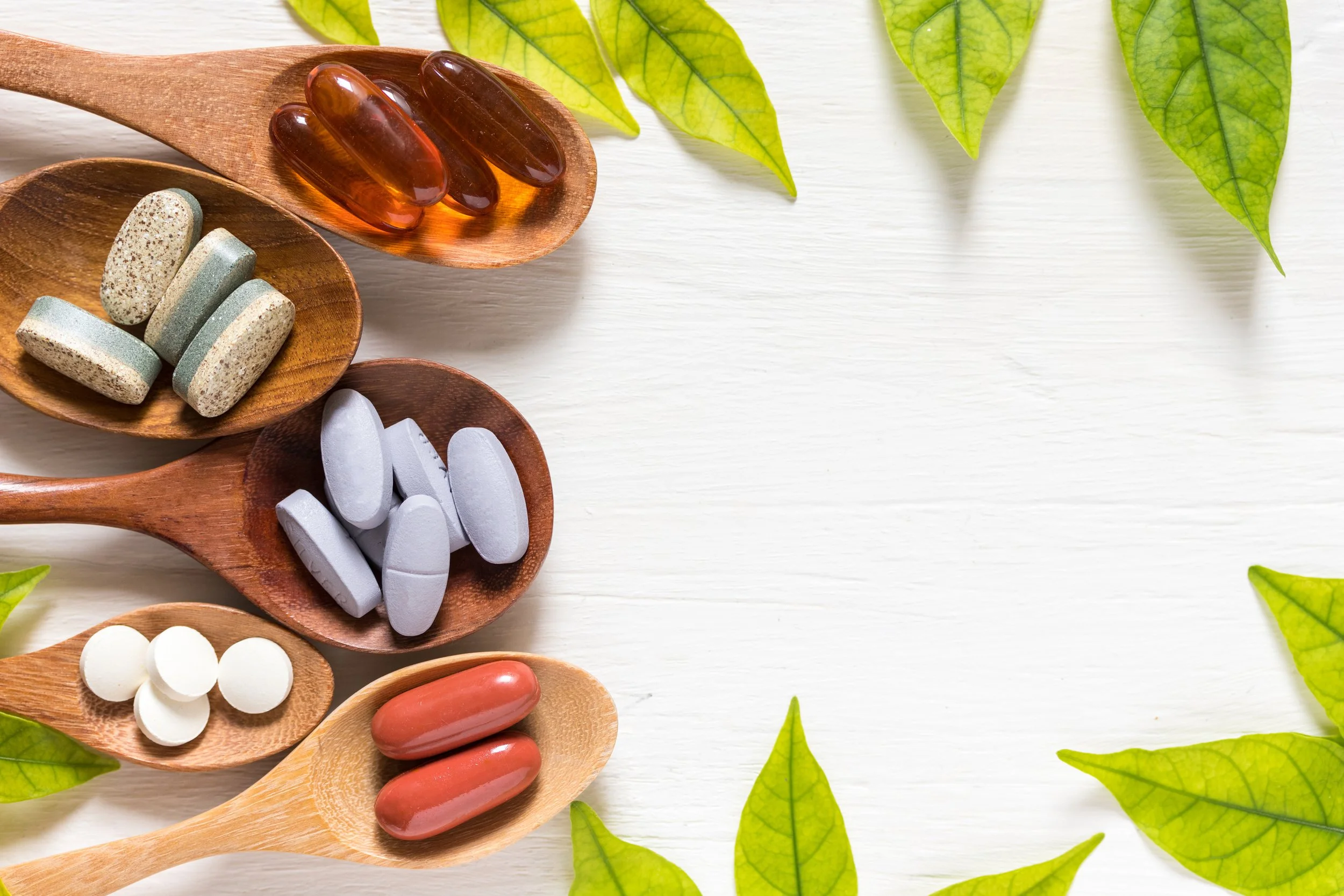 Various pills and capsules on wooden spoons and dishes arranged on a white surface with green leaves around the edges.