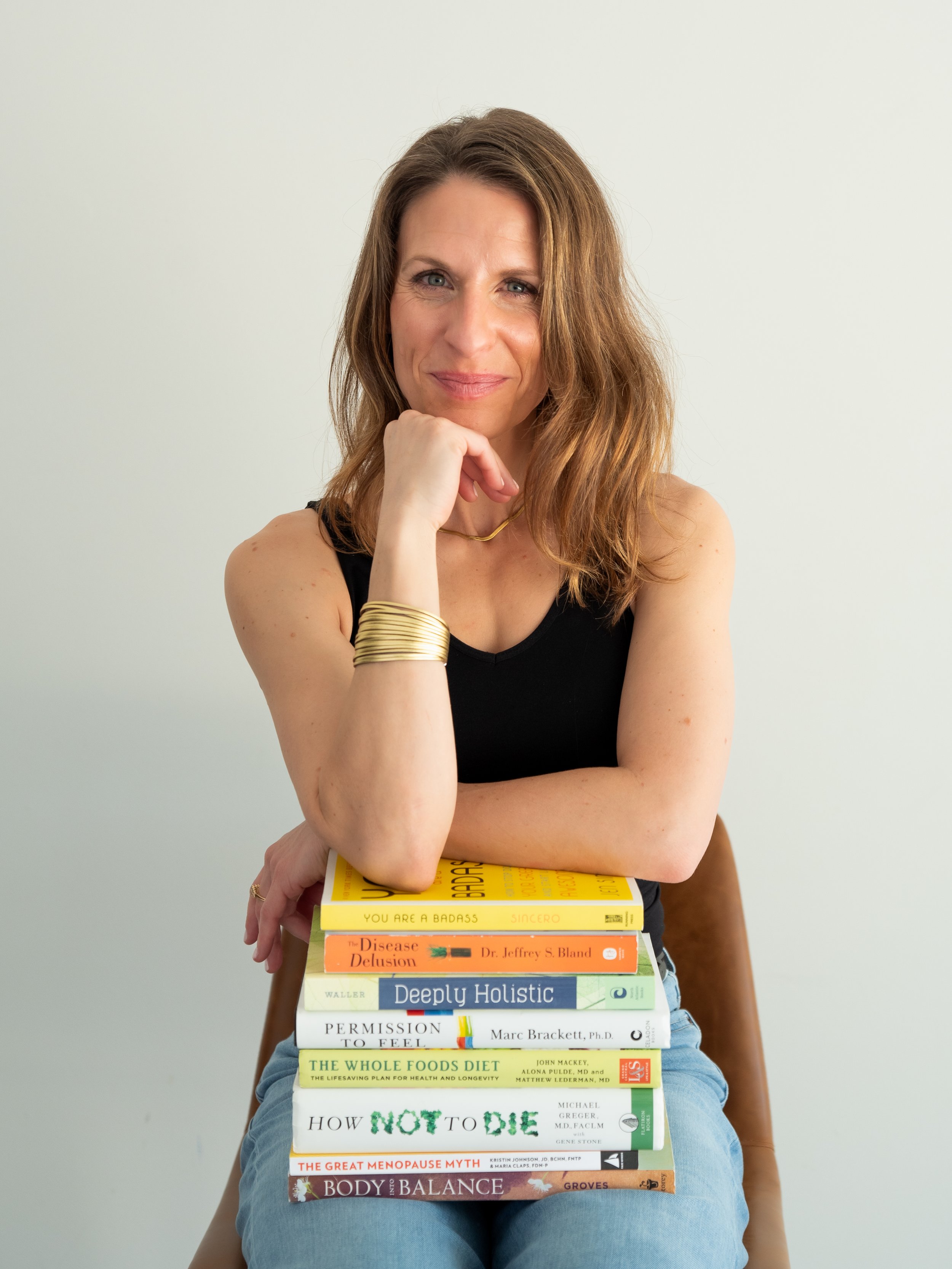 A woman with light brown hair and blue eyes, wearing a black top and gold jewelry, sitting and holding a stack of books in her lap. Her right hand rests on her chin.