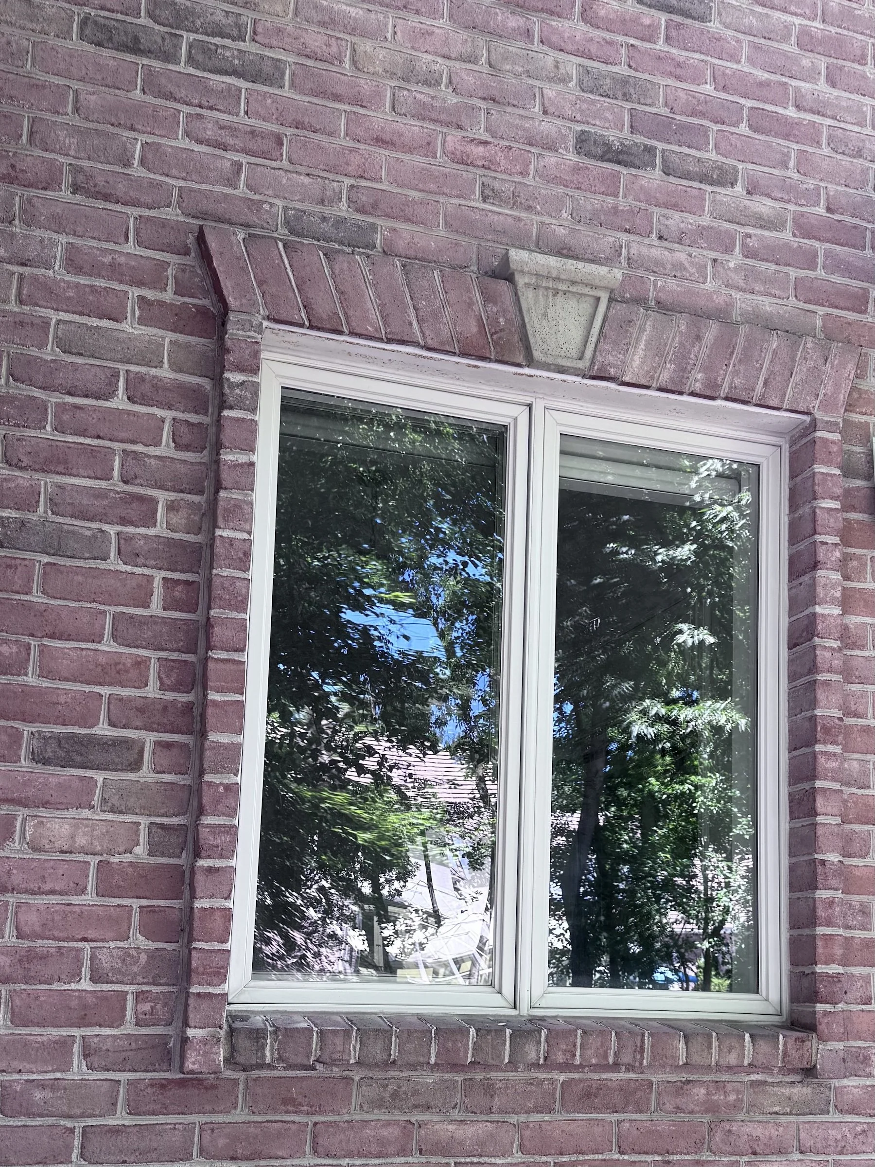 A brick wall with a white-framed window reflecting green trees and a blue sky, with a concrete outdoor light fixture above the window.