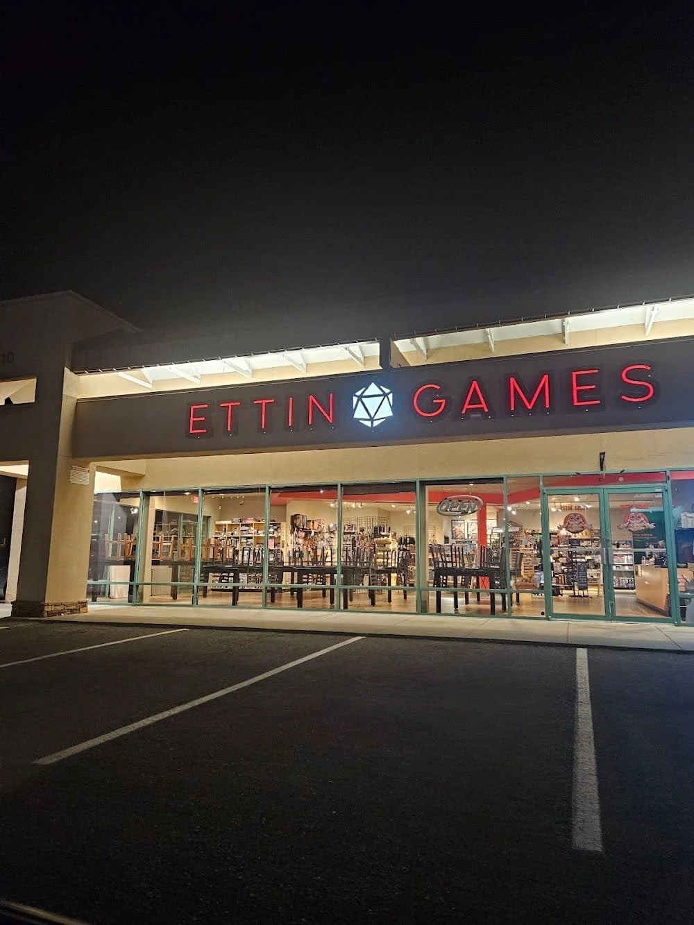 Night view of the exterior of Ettin Games store with large glass windows, illuminated sign, and empty parking lot in front.
