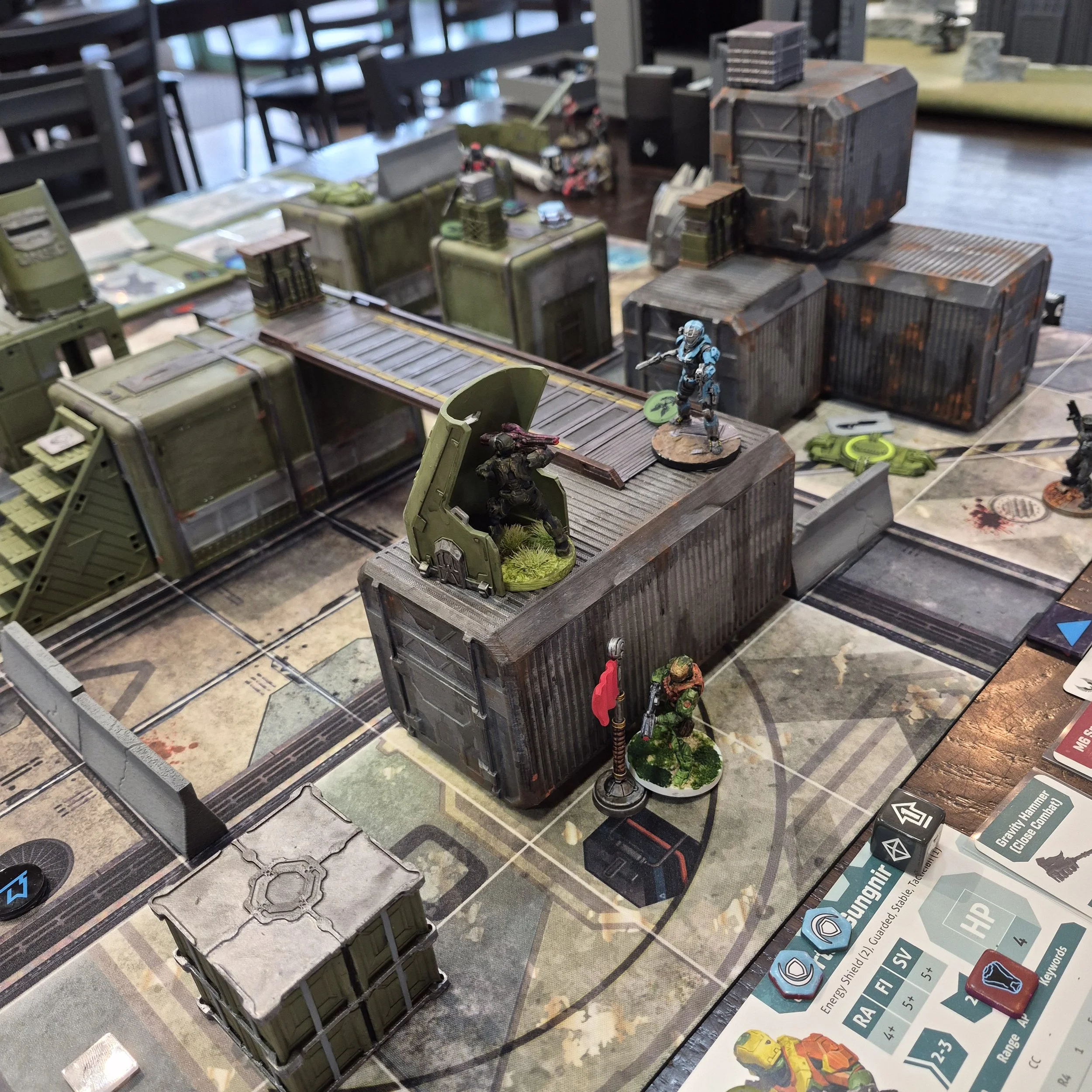 A tabletop wargame setup with miniature soldiers, vehicles, and buildings, including a blue armored figure and various terrain pieces.