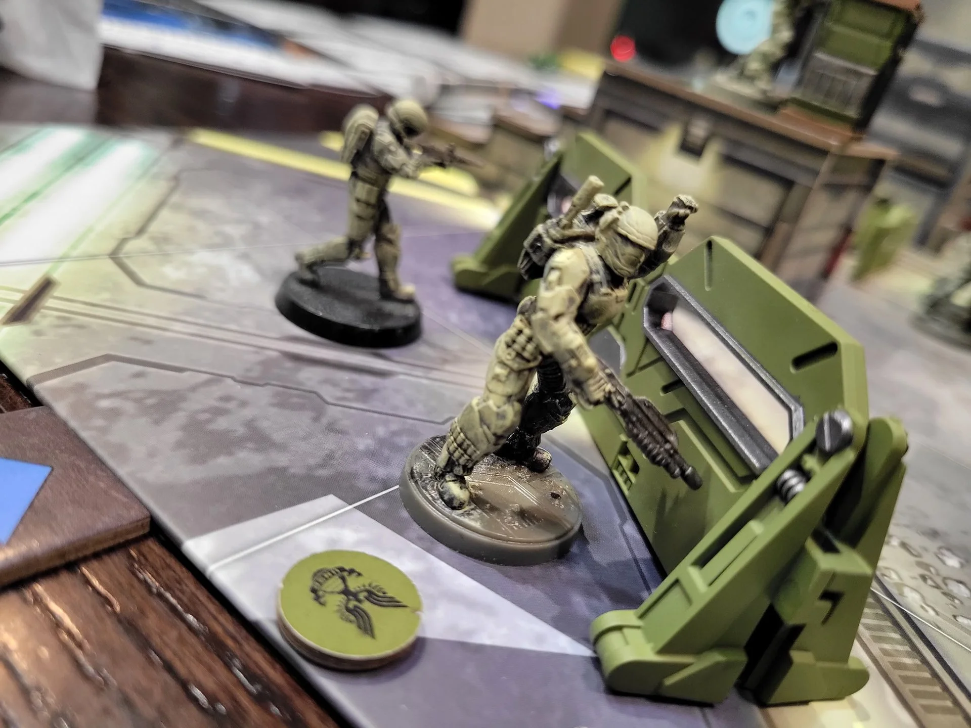 Tabletop gaming scene with two military miniature figures, one in beige camouflage and the other in darker camouflage, positioned behind green gaming accessories and a game board.