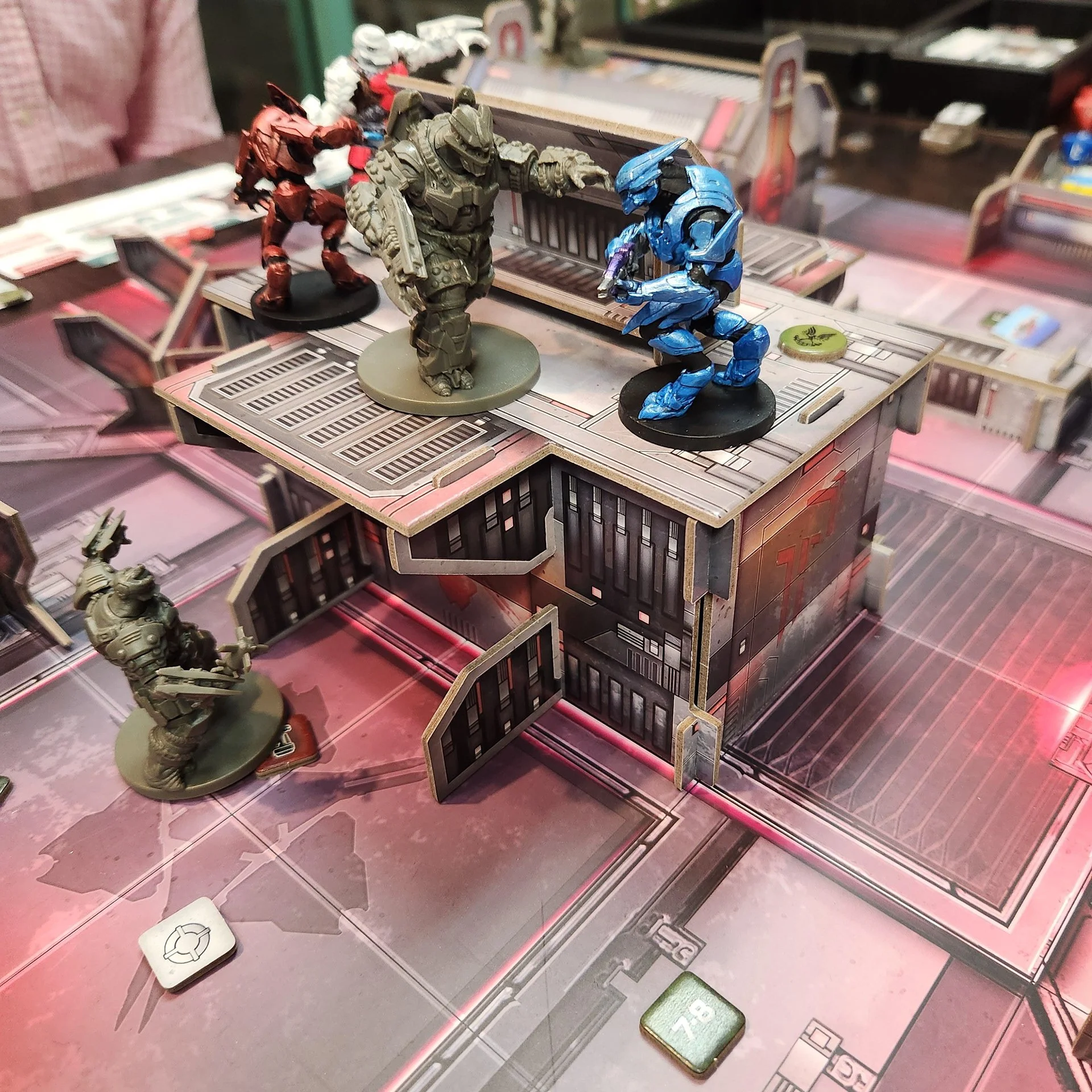 Three miniature figures from a sci-fi tabletop game are positioned on a detailed game board with a futuristic cityscape layout. One figure is red, another is gray, and the third is blue, with the blue figure kneeling and pointing at the gray figure, 