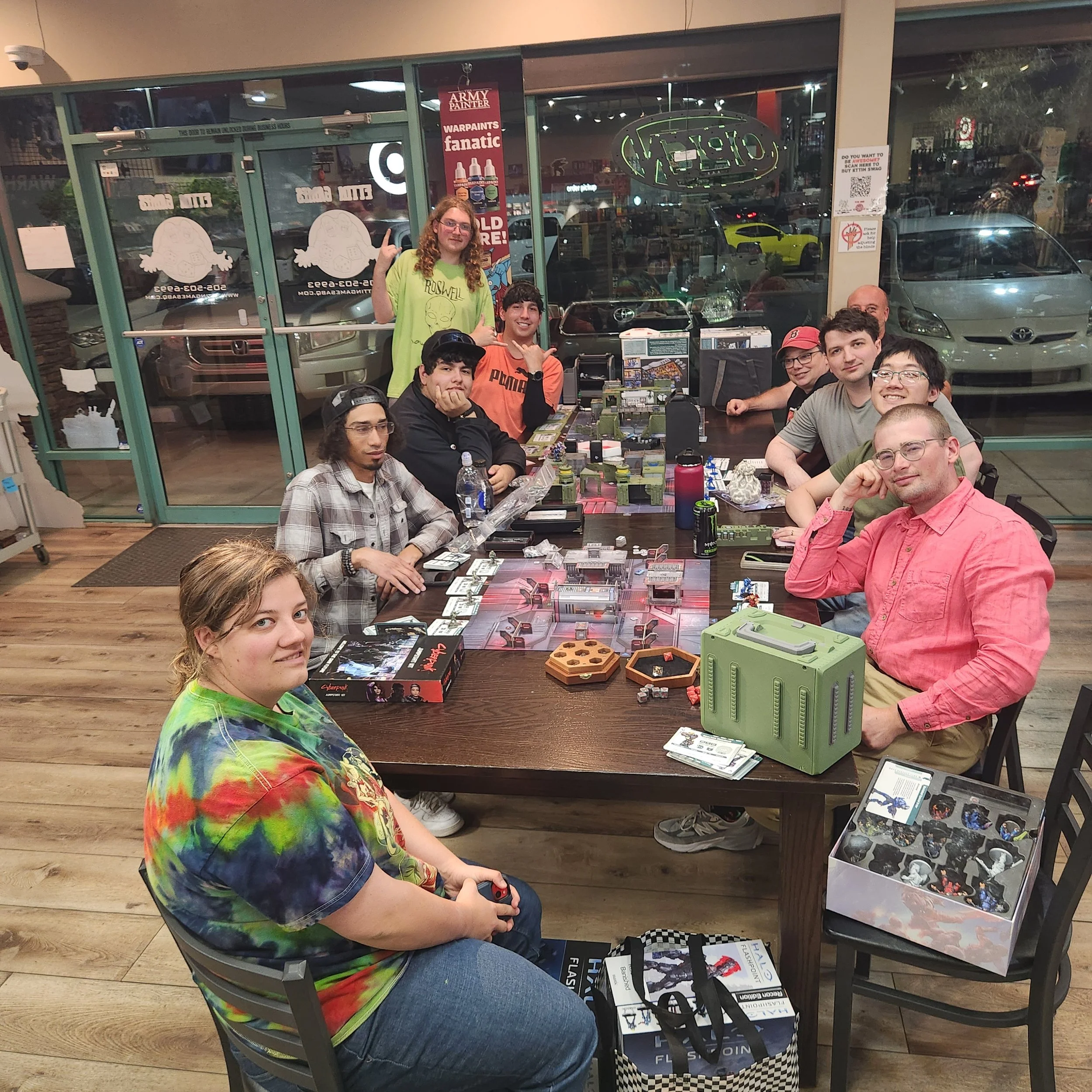 A group of people sitting and standing around a table with tabletop gaming equipment inside a store. The store has large glass windows and doors with signs and advertisements. A young woman in a colorful tie-dye shirt is sitting at the front, and a young man in an orange shirt next to her is making a shaka hand gesture. Multiple other individuals are seated around the table, smiling and engaging with the gaming materials.