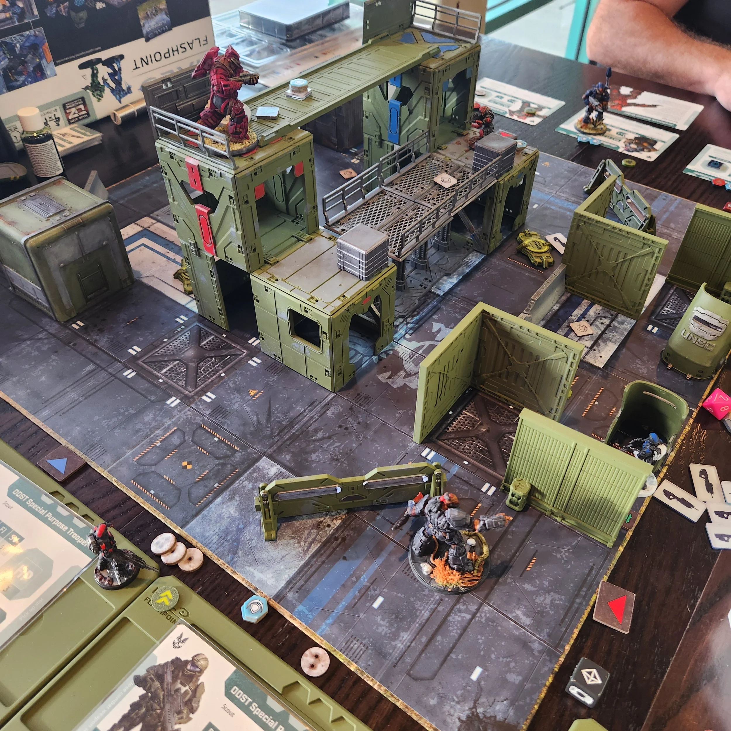 Tabletop wargame in progress with miniature soldiers, vehicles, and terrain pieces, including a large green military-style building and scattered game cards and tokens.