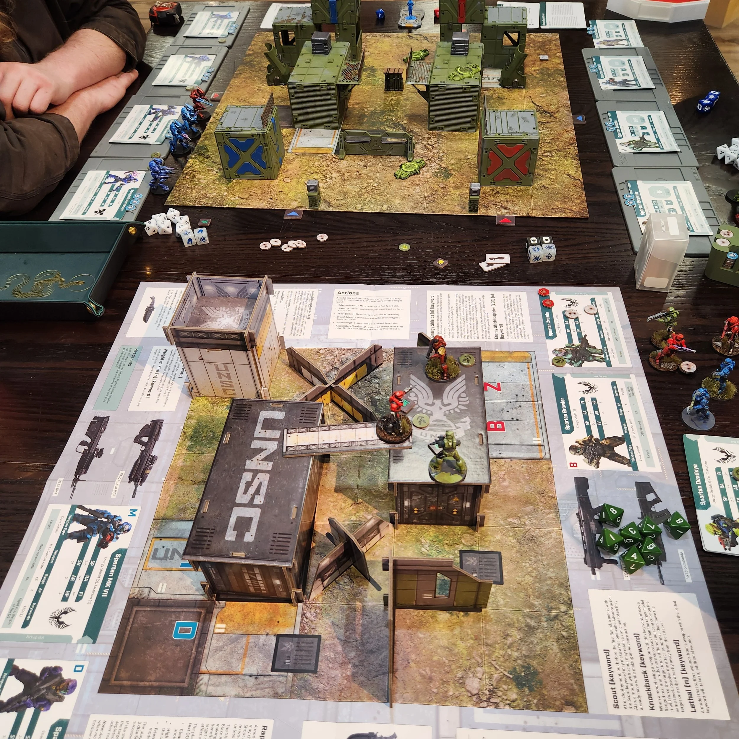Tabletop miniatures game with buildings, miniature soldiers, dice, and game sheets on a table.
