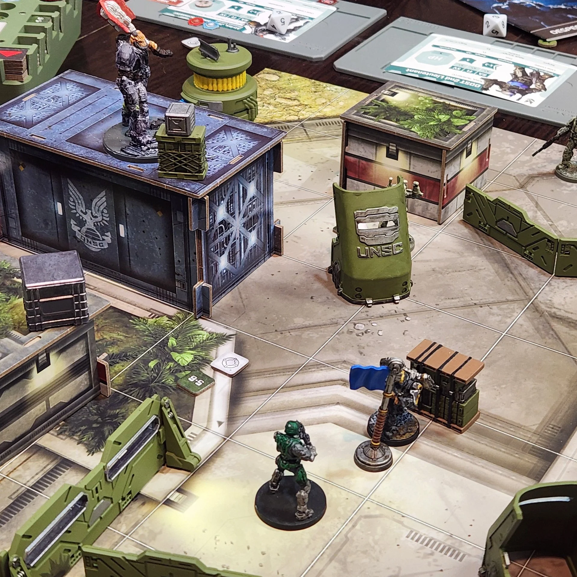 Tabletop gaming setup with miniature soldiers, terrain pieces, game cards, and dice, arranged on a tiled floor.