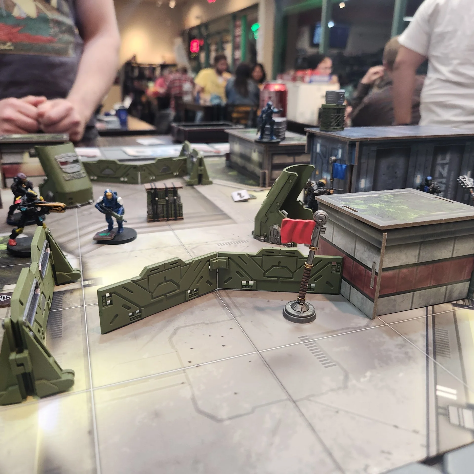 Tabletop with miniature figurines and green terrain pieces, set up for a tabletop game in a gaming store or arcade, with people in the background.