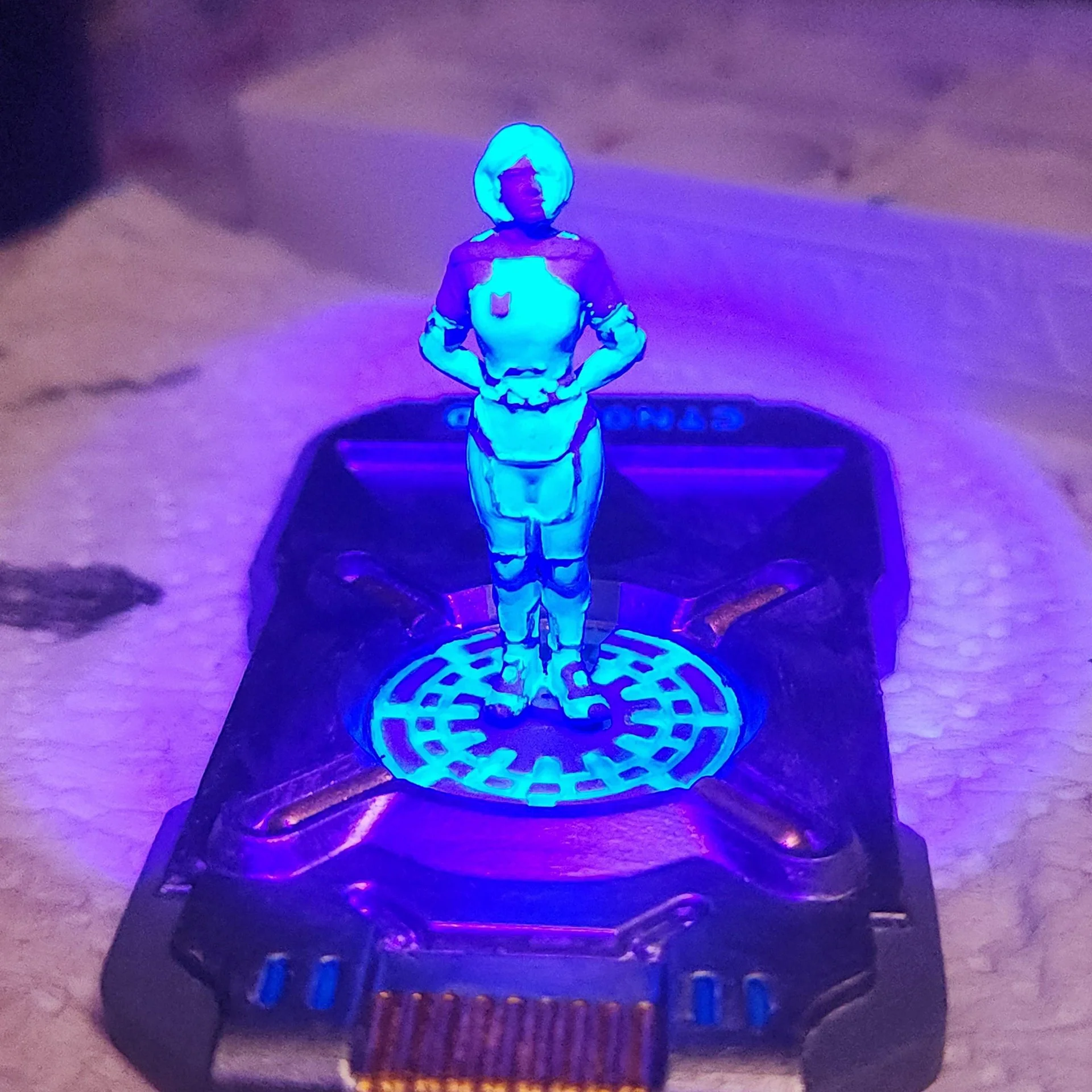 A small black plastic device with a blue illuminated decoration on top, featuring a figurine of a person in a superhero or armor costume, with a luminous blue halo around the head, positioned on a flat surface.