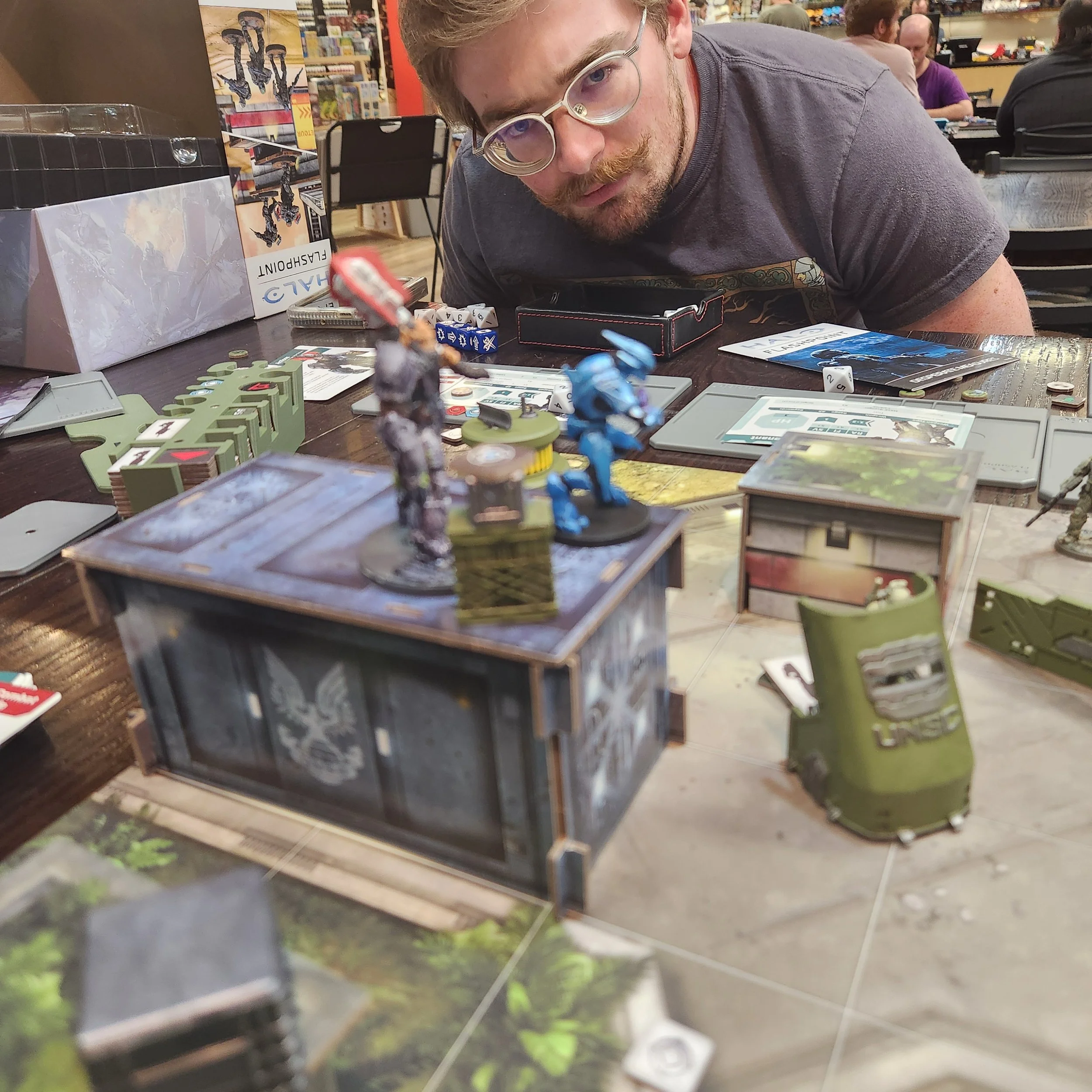 A person with curly hair and glasses leaning over a table with figurines, game cards, and dice in a store or gaming cafe.