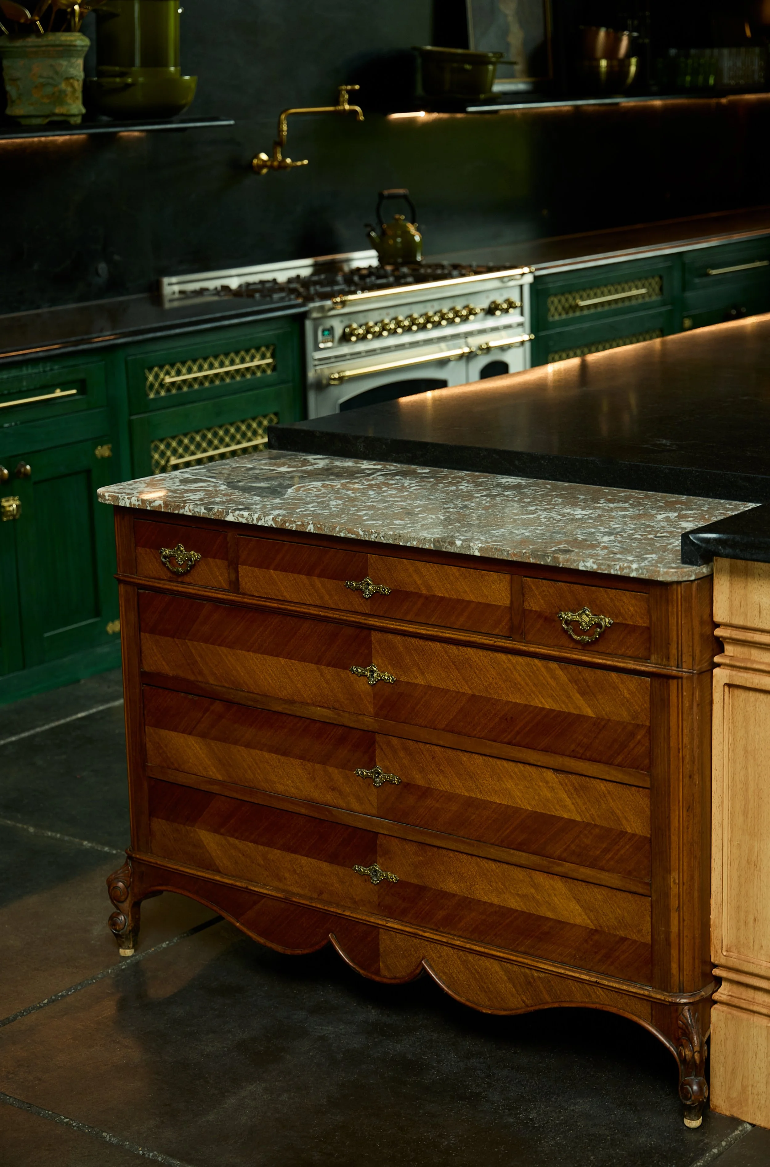 An antique wooden dresser with a marble top and gold ornate handles, placed in front of a dark kitchen with green cabinetry and a vintage stove, on a dark floor.