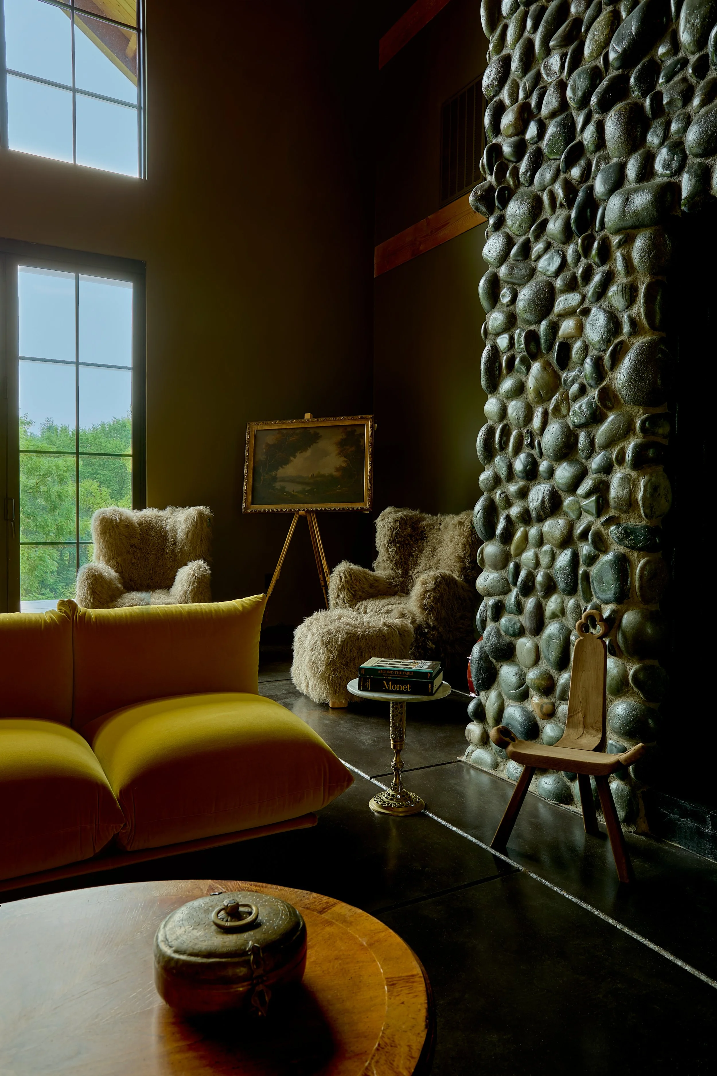 Interior of a cozy living room with a yellow velvet sofa, a round wooden coffee table with a decorative jar, a furry armchair, a framed landscape painting on an easel, and a large stone wall. There are tall windows with a view of green trees outside.