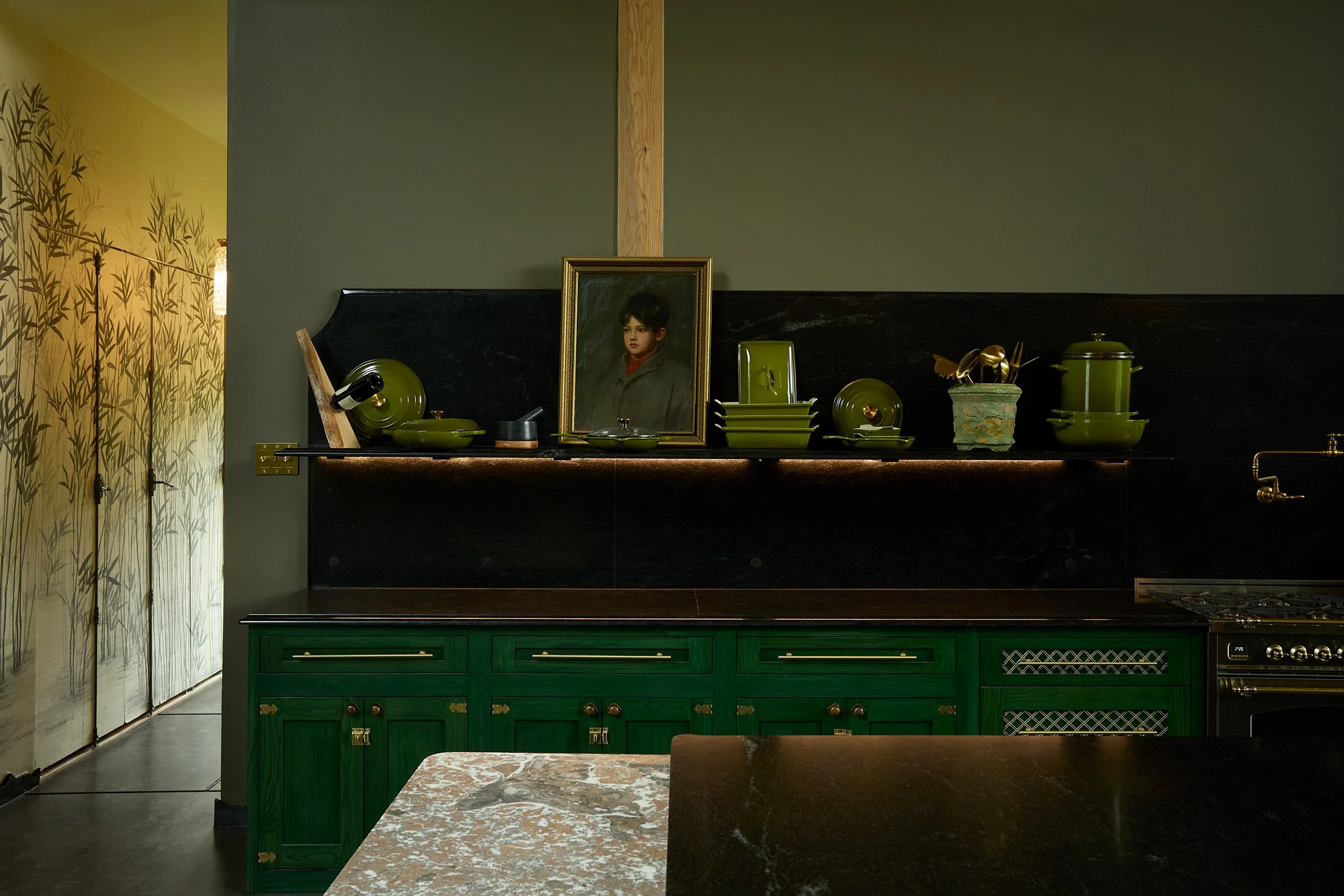 A kitchen with a dark marble countertop and green cabinets, decorated with a framed portrait of a young boy, green dishware, and a decorative pottery item, with a yellow floral wallpaper on the left side.
