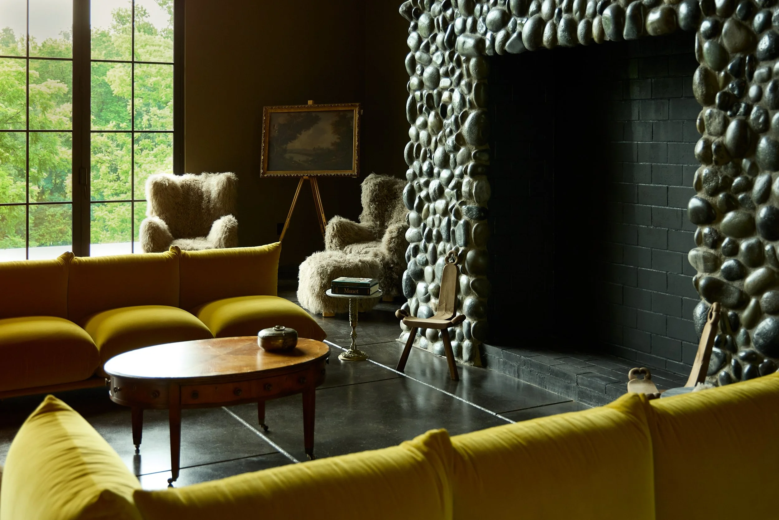 A cozy living room with large windows showing green trees outside. The room features a yellow sofa, two fluffy armchairs, a wooden coffee table, a framed landscape painting on an easel, and a stone fireplace with a dark brick interior.