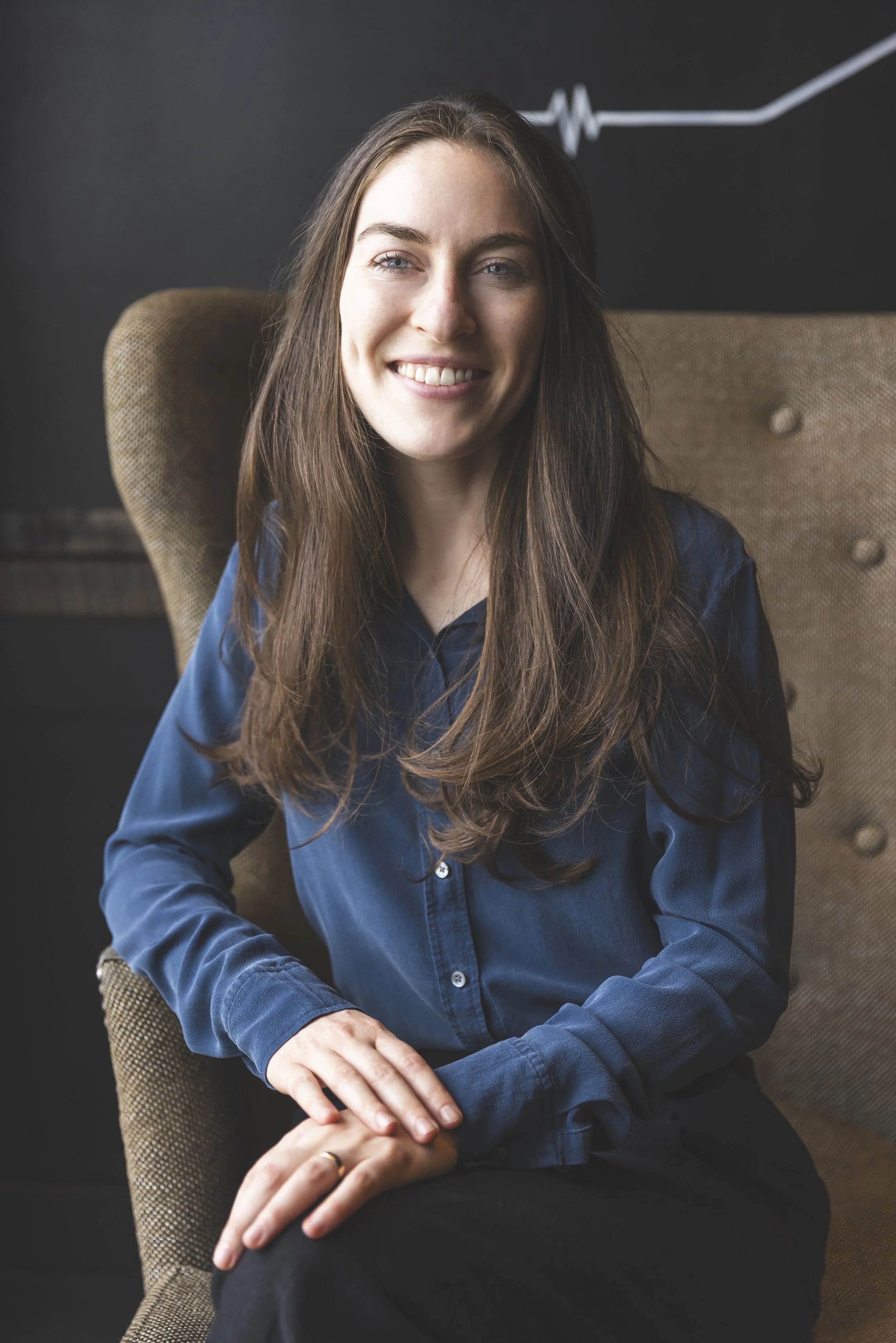 A smiling woman with long brown hair, wearing a blue shirt, sitting in a brown upholstered chair against a dark background.
