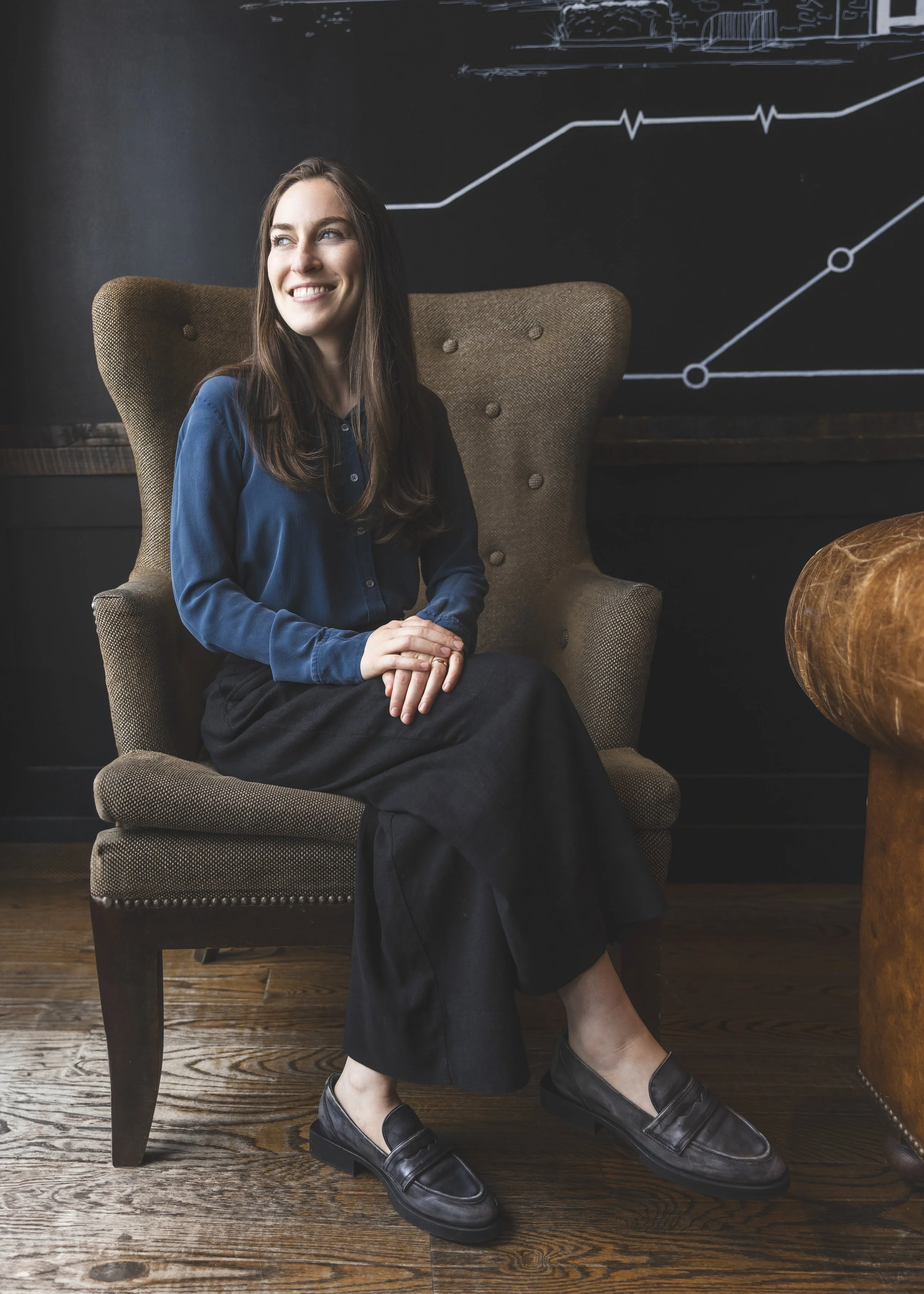 A woman with long brown hair and a smile, wearing a blue blouse, black wide-leg pants, and black loafers, sitting in a vintage armchair in a room with dark walls and a wooden floor, with white line diagrams on the wall behind her.